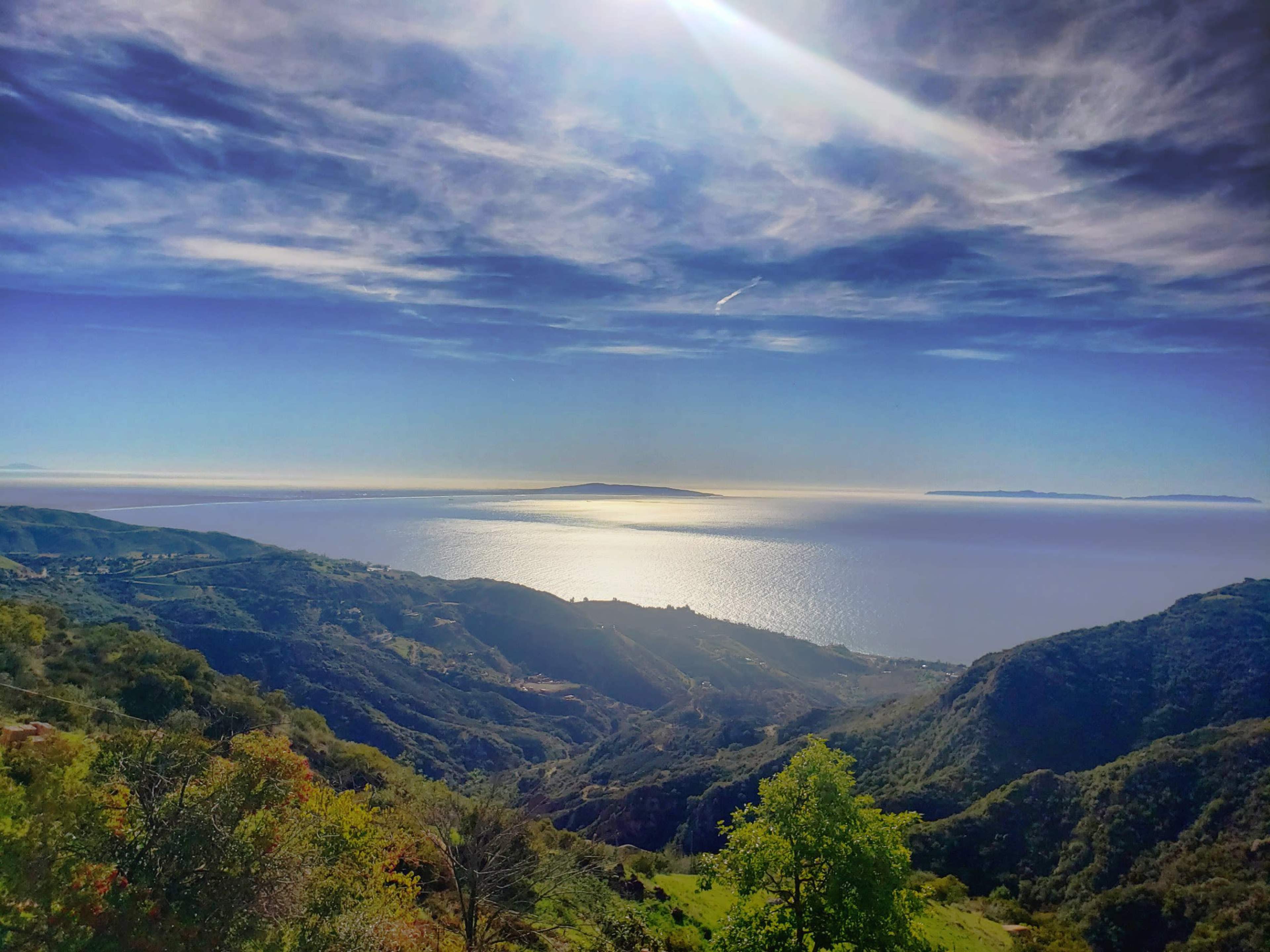 The image showcases a panoramic view of a coastal landscape with rolling hills descending to the sea, reflecting sunlight under a clear blue sky.