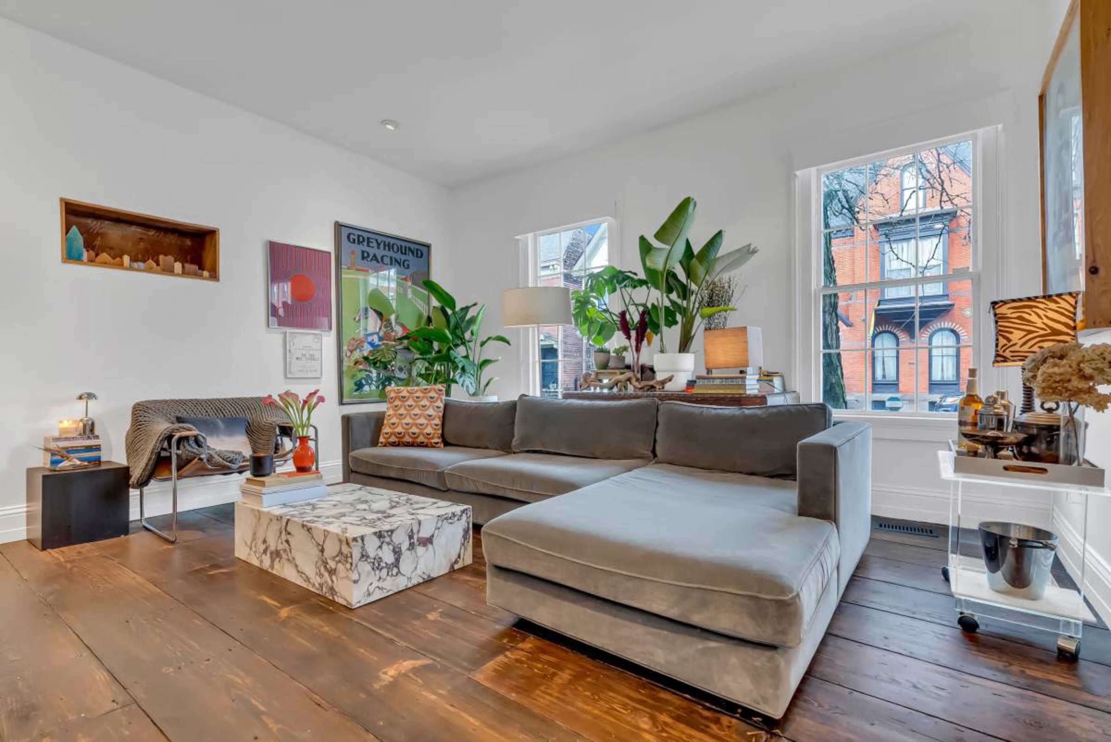 The living room features a gray sectional sofa, a marble-topped coffee table, and several indoor plants, with large windows allowing natural light.