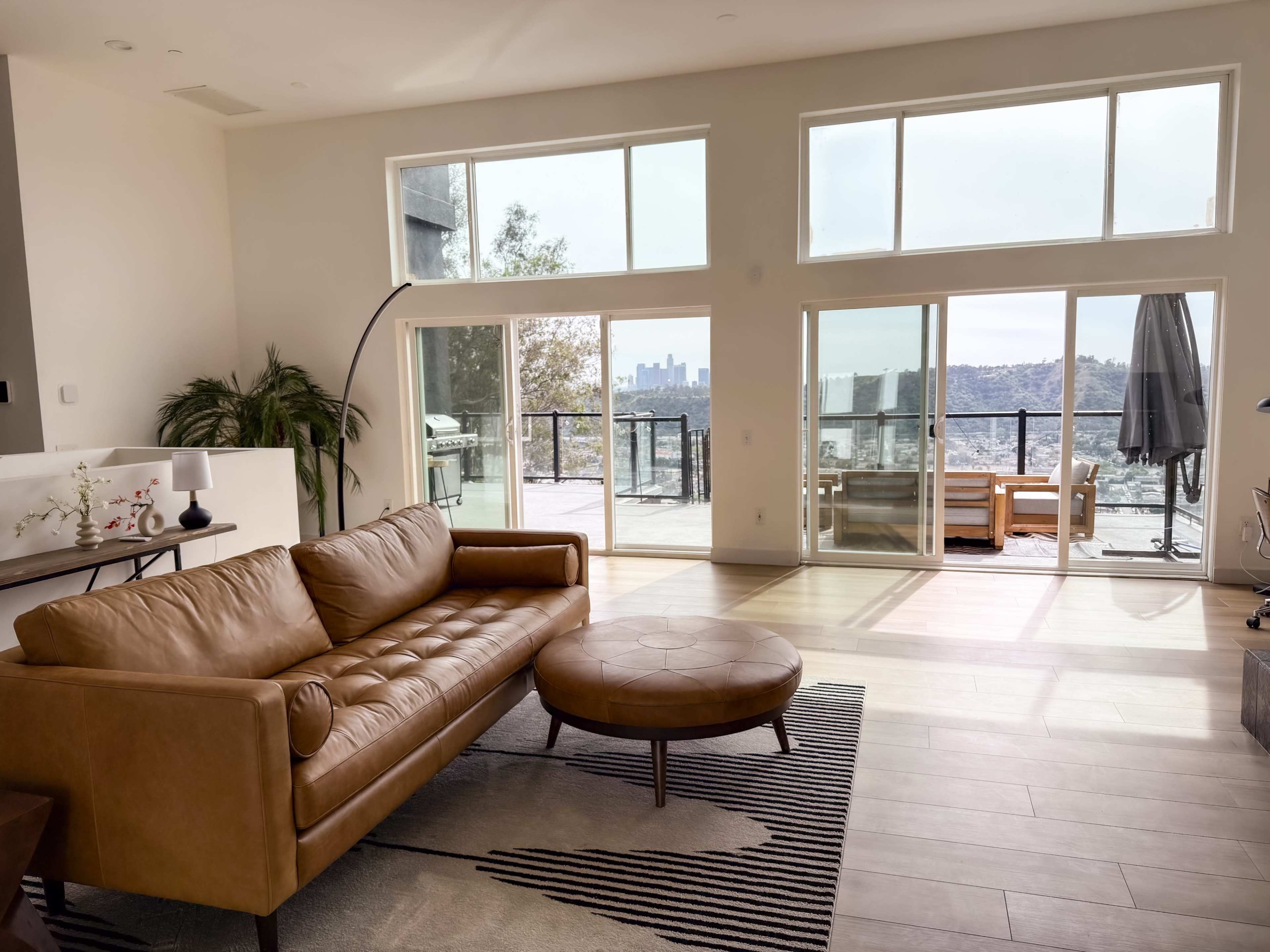 The image shows a modern living room with a brown leather sofa and an ottoman, featuring large windows that overlook a city skyline and a terrace.