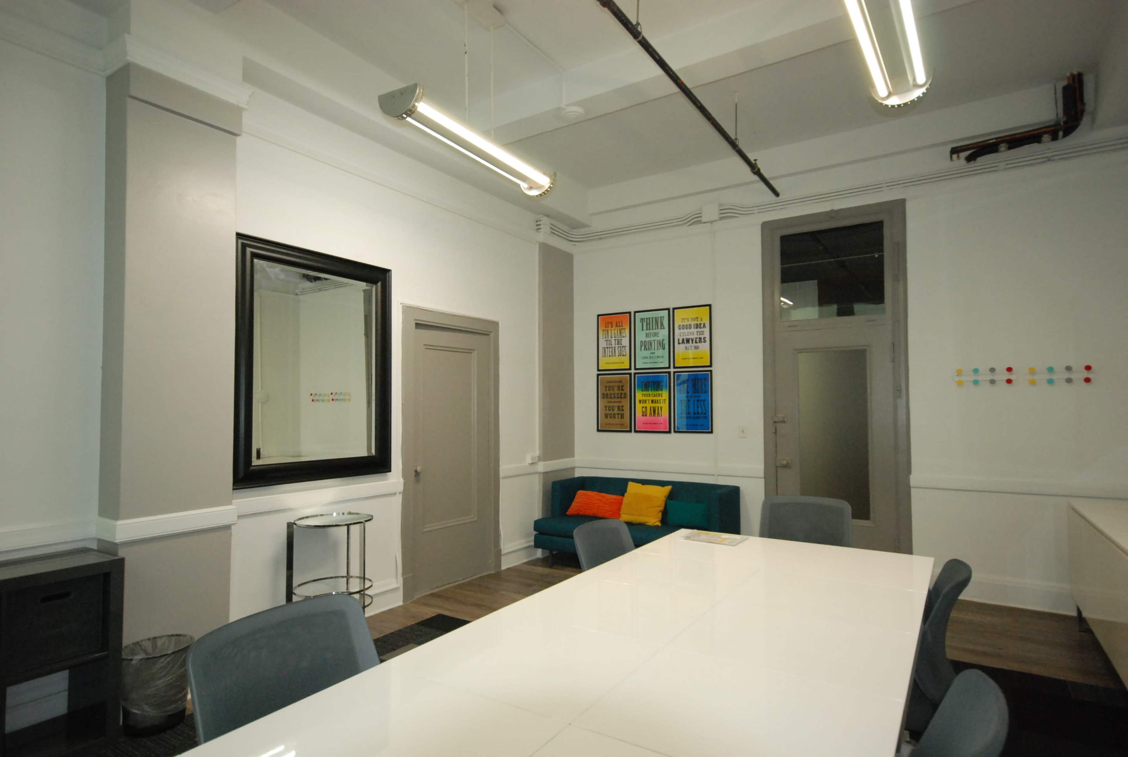 The image shows a modern conference room with a large white table, a green couch with colorful cushions, framed posters on the wall, and a mirror reflecting part of the room.