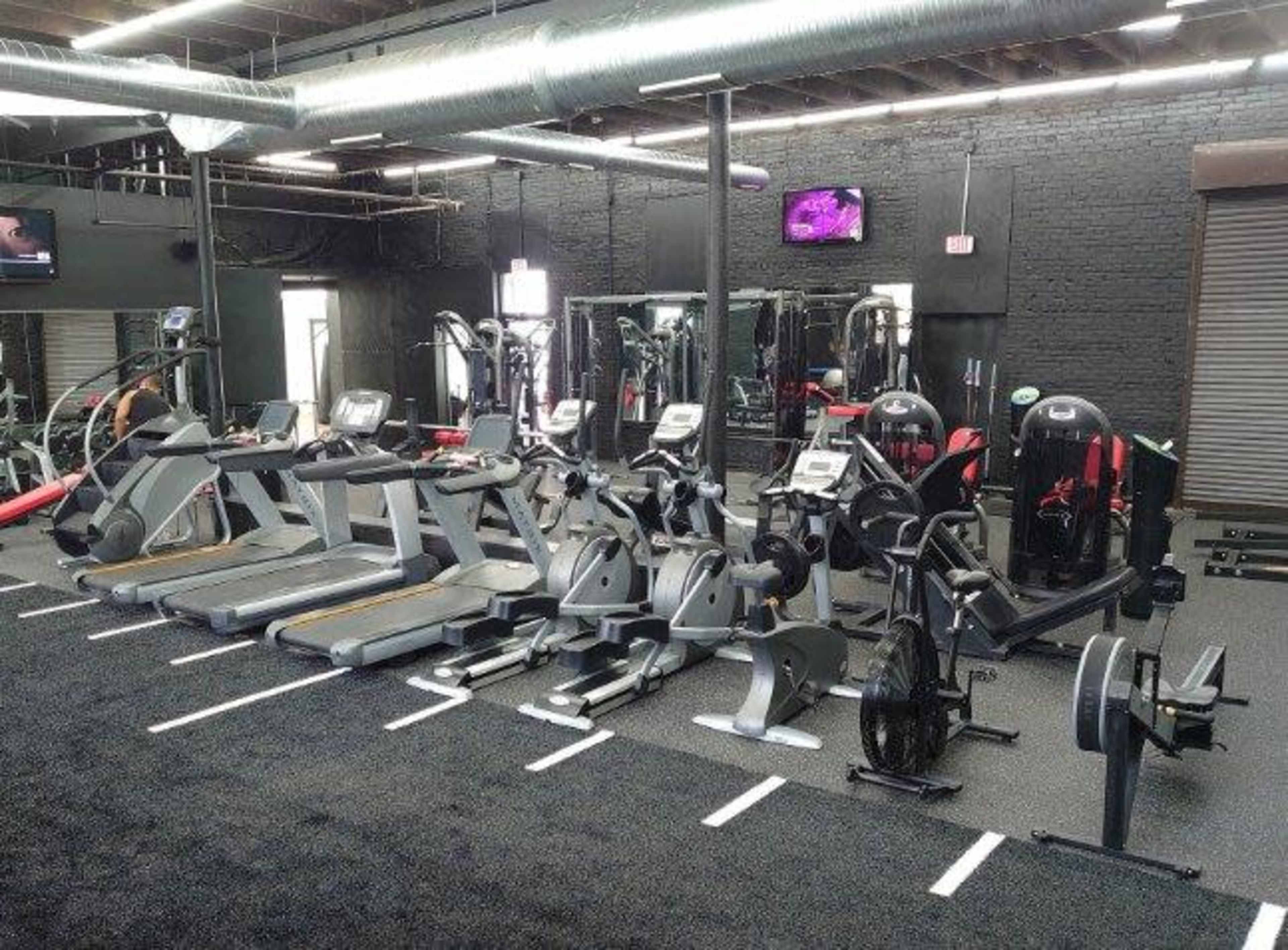 The image shows a gym interior with various fitness machines arranged neatly on a black rubber floor.