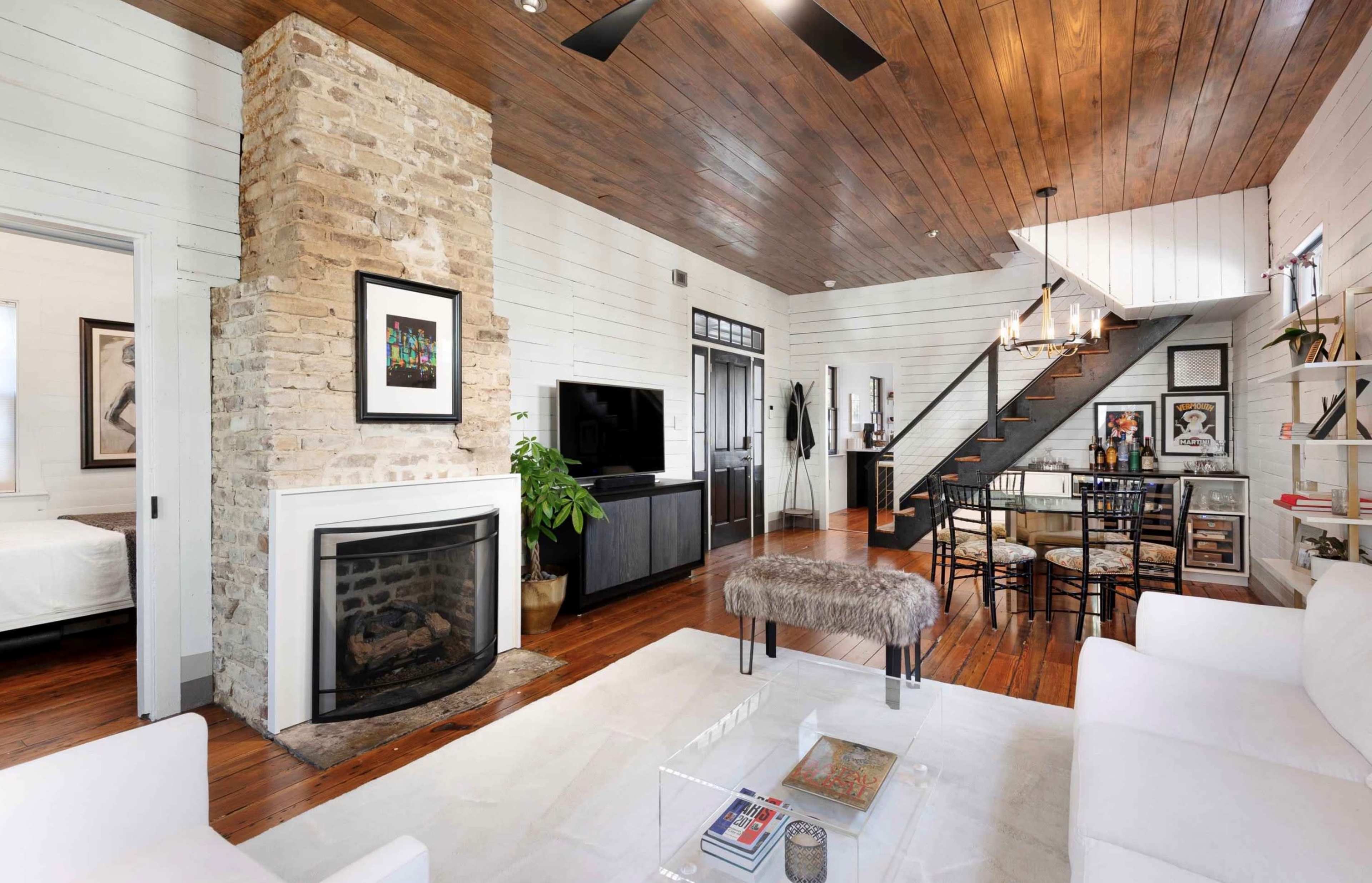 A modern living area with a stone fireplace, wooden ceiling, and open staircase leading to a loft space, furnished with a white sofa and a dining table.