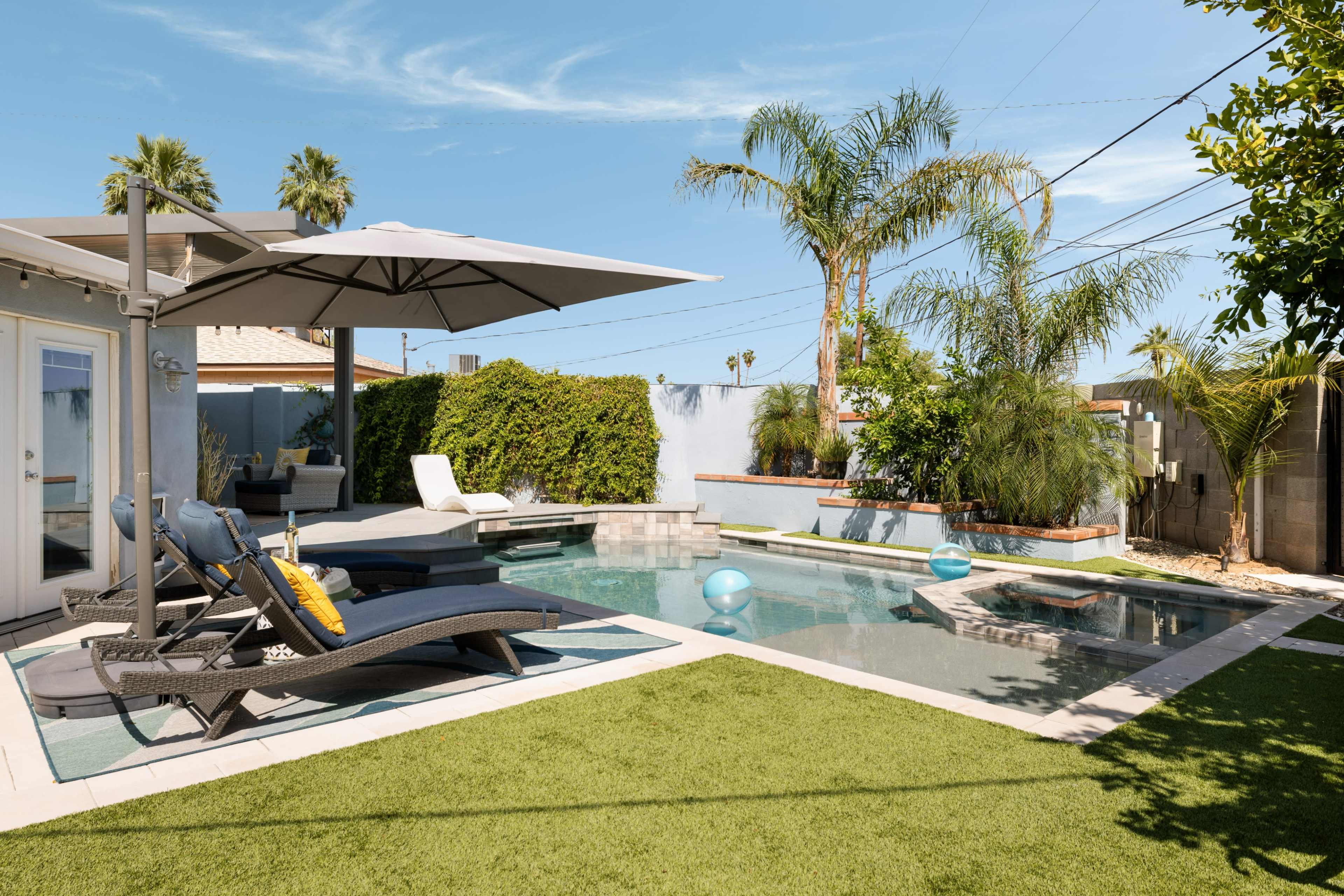 The image shows a backyard pool area with lounge chairs, a large umbrella, and tropical plants surrounding a clear swimming pool.