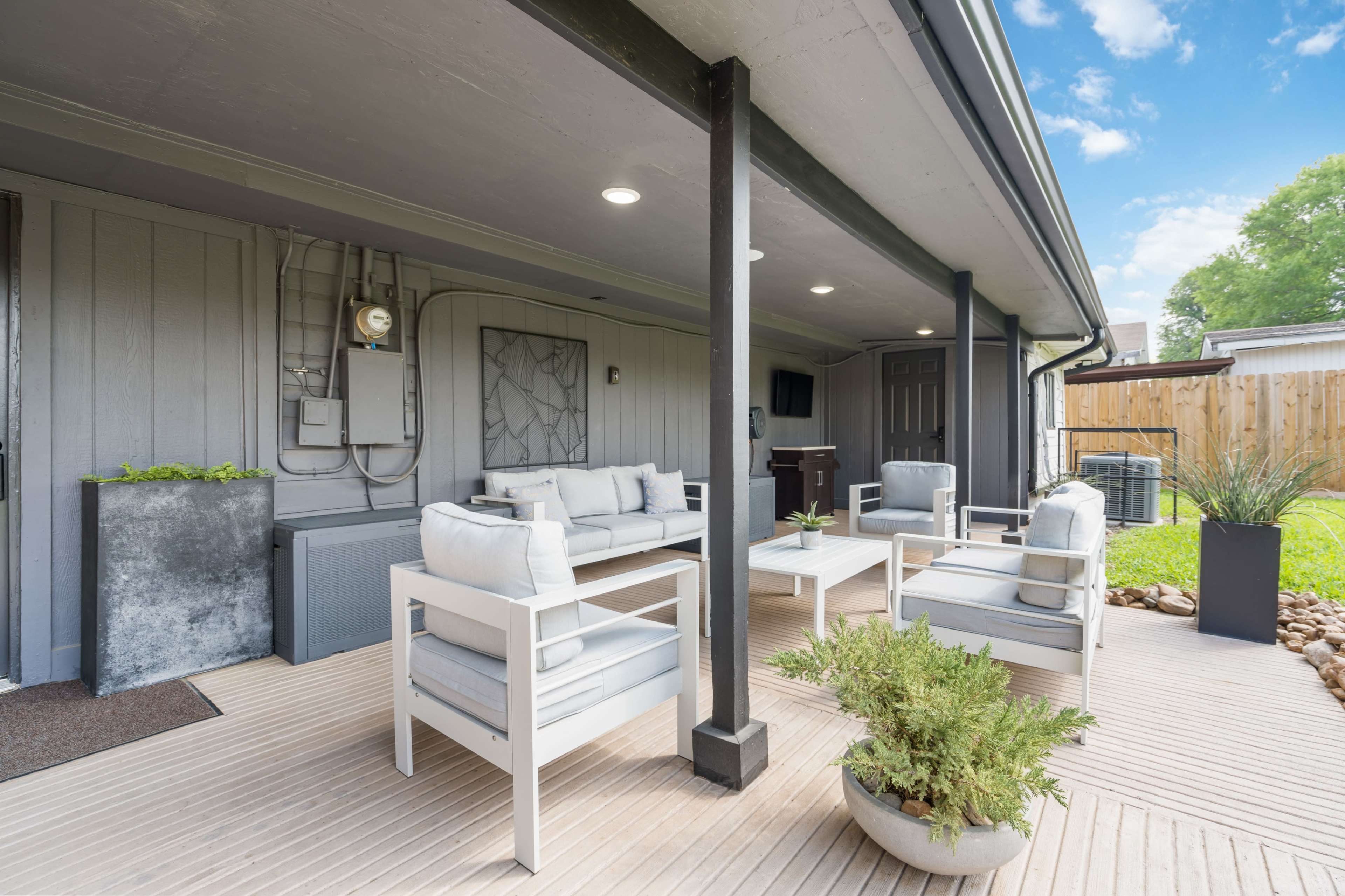 A modern outdoor patio area with white cushioned seating, a small table, and decorative plants, set against a wooden deck and an exterior wall.