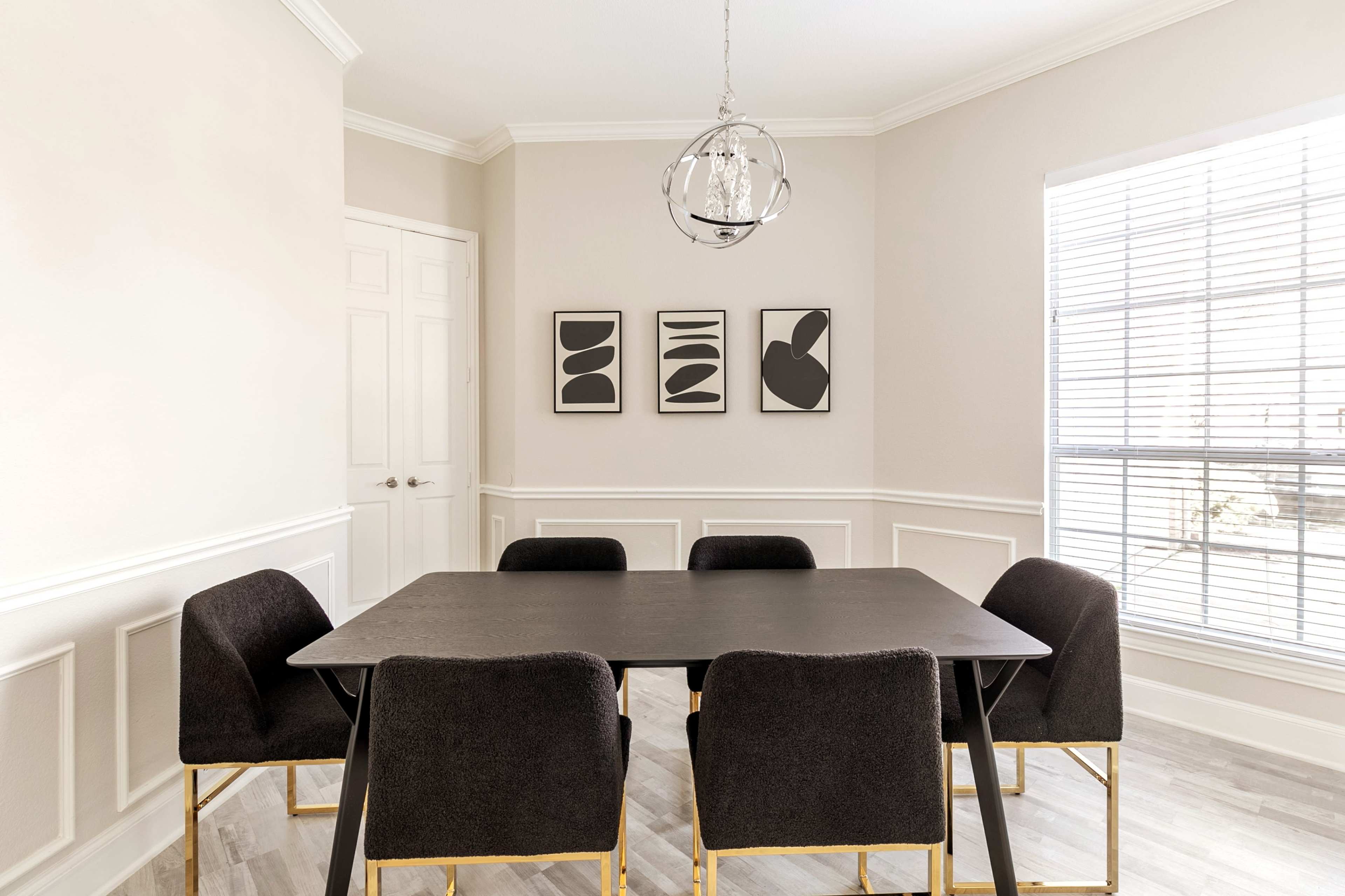 A dining room features a black table surrounded by four dark upholstered chairs, with three abstract art pieces on the wall and a window allowing natural light.