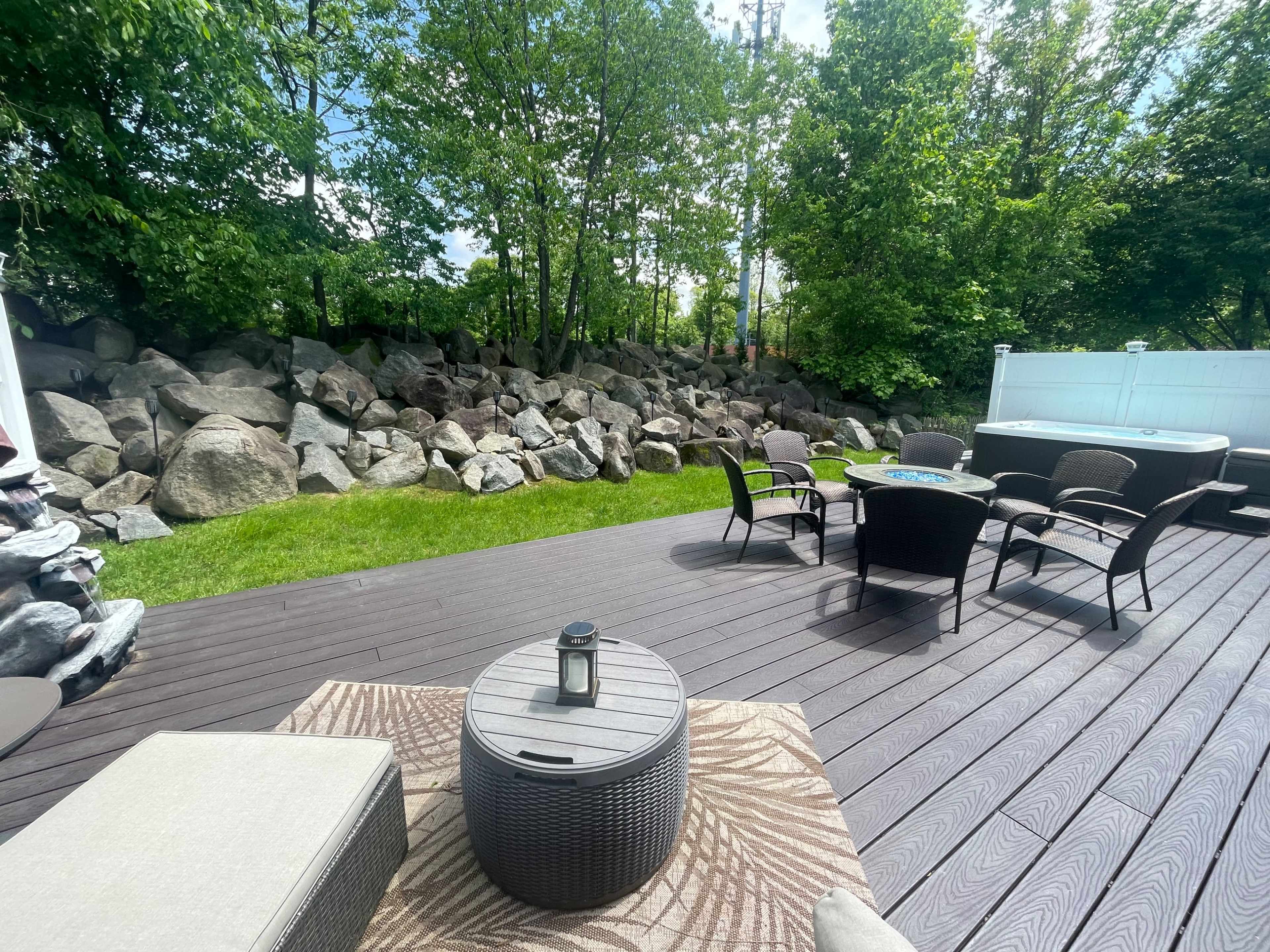 A backyard patio with outdoor furniture, a round table, and a rock wall surrounded by greenery.