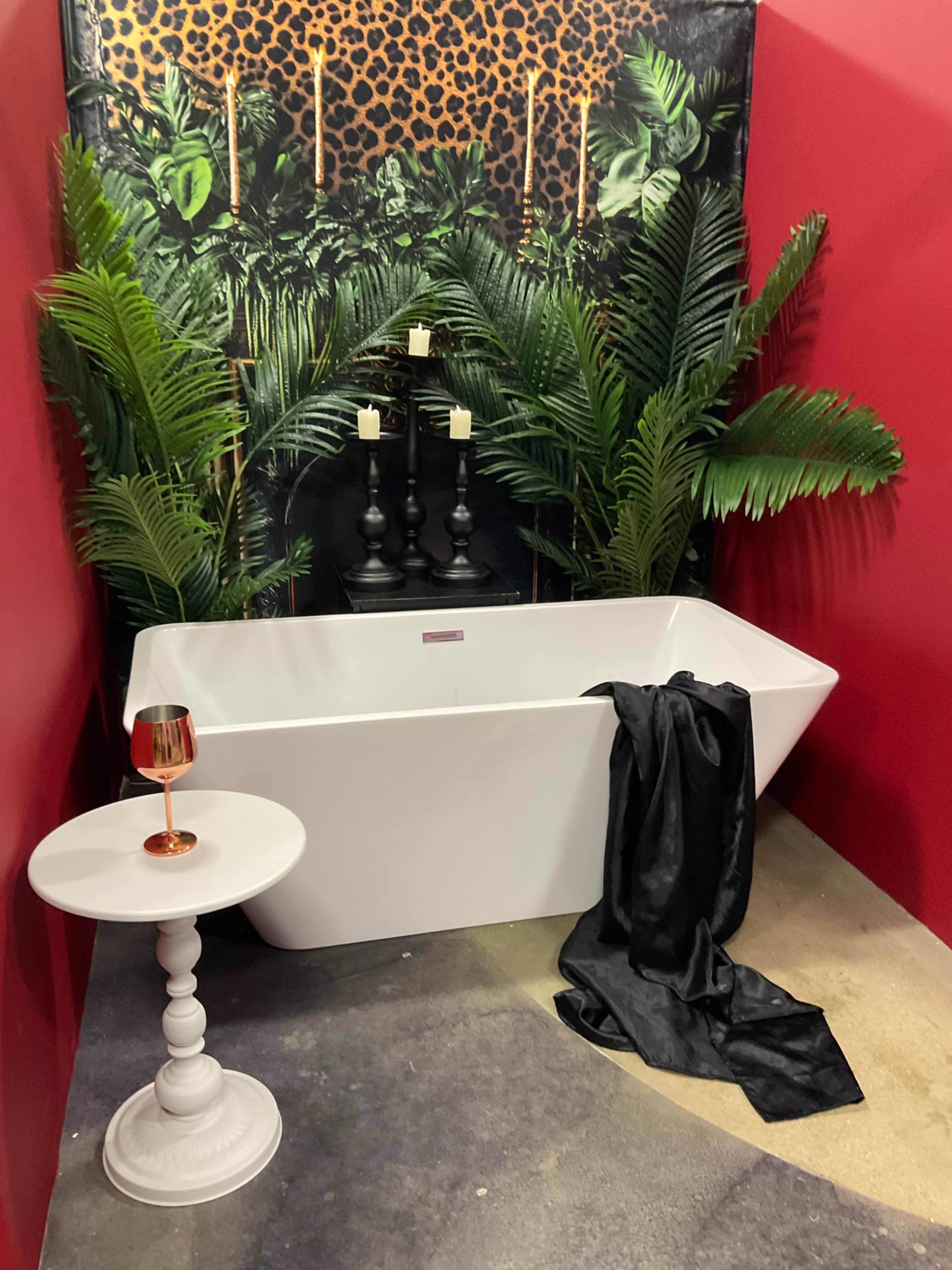 A modern white bathtub is positioned in front of a leopard-print wall, surrounded by tall green plants and accompanied by a small white side table with a decorative candle.