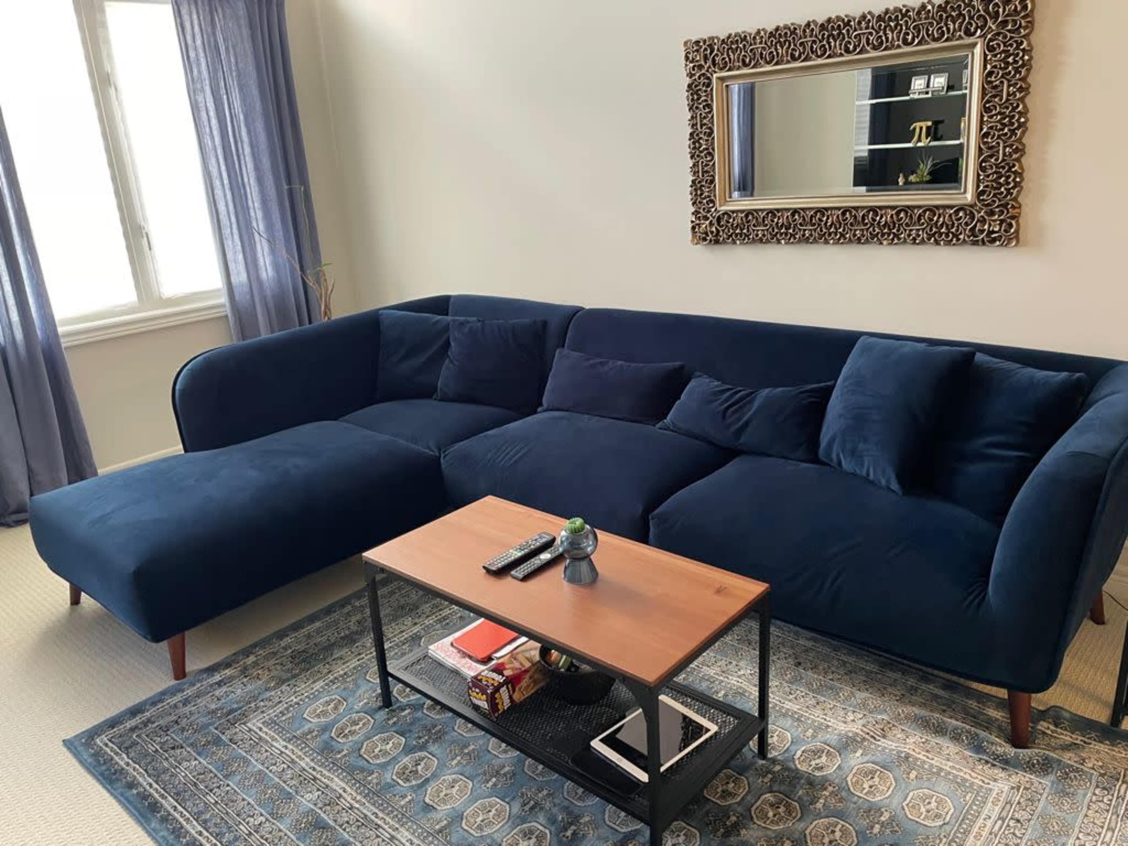 A blue sectional sofa with cushions sits beside a wooden coffee table on a patterned rug in a well-lit living room.