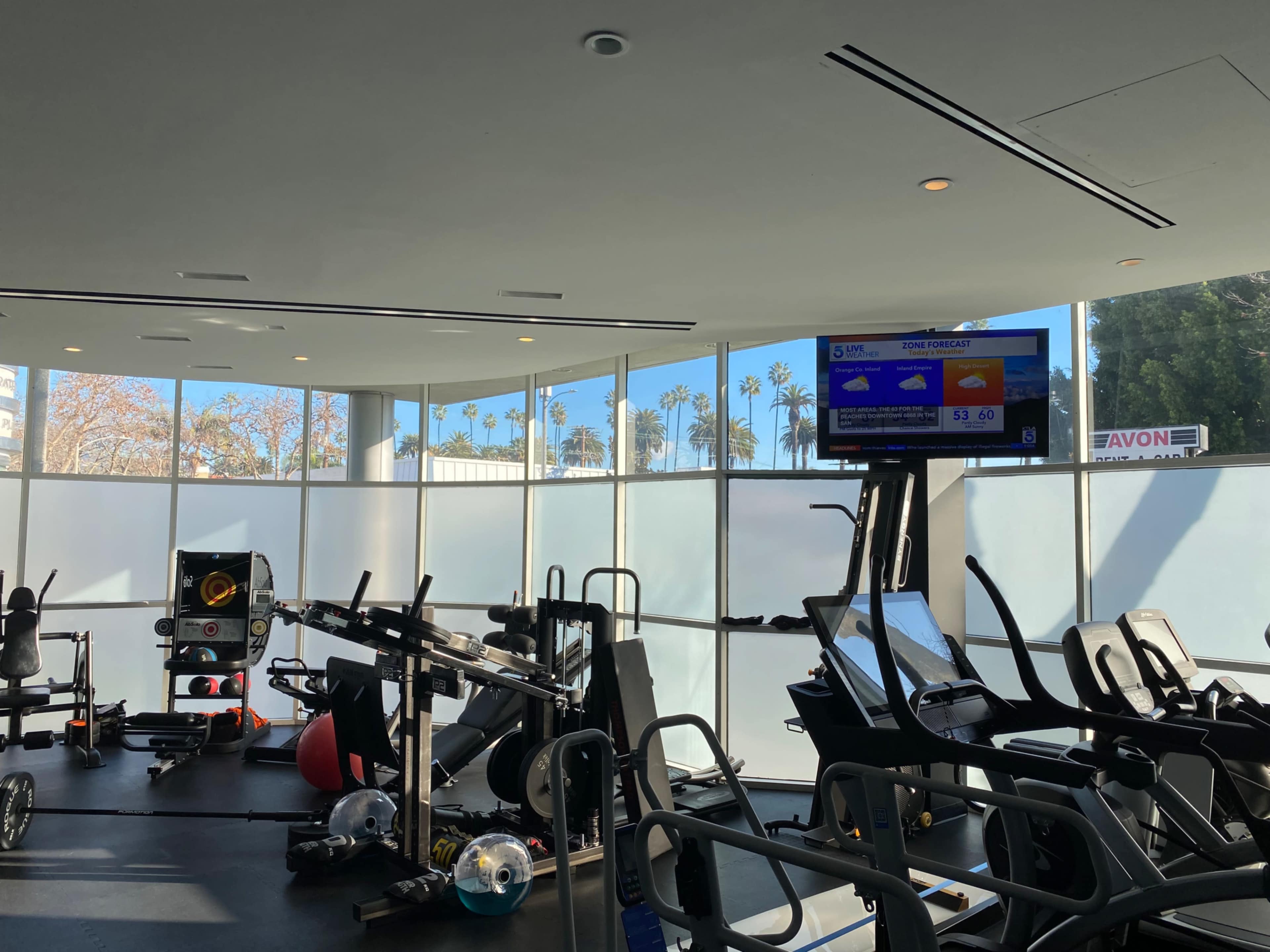 The image shows a modern gym with exercise equipment and a large screen displaying weather information, illuminated by natural light coming through large windows.