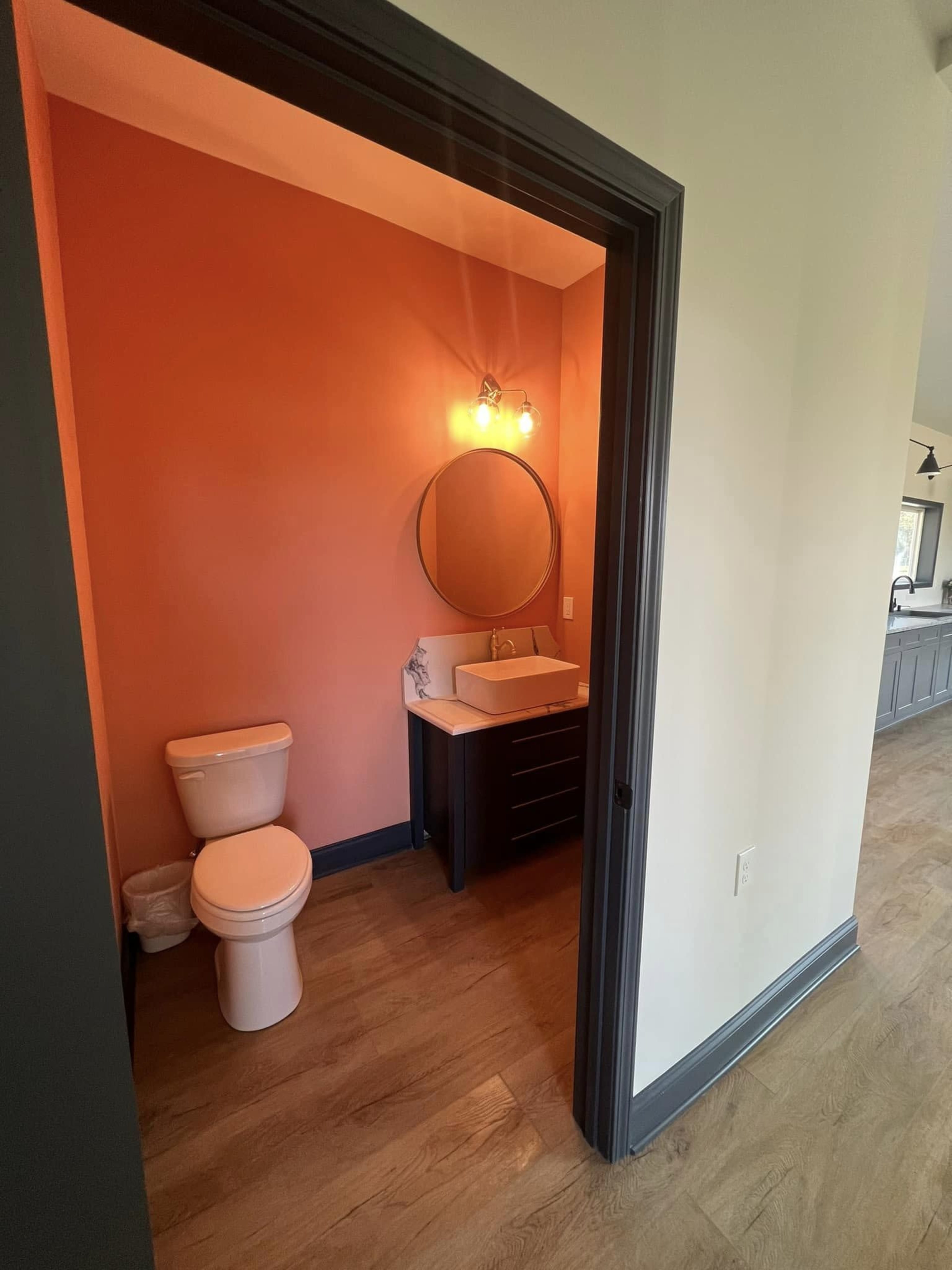 The image shows a small bathroom with a round mirror and a sink unit, featuring a toilet and light-colored walls contrasting with the darker framing.