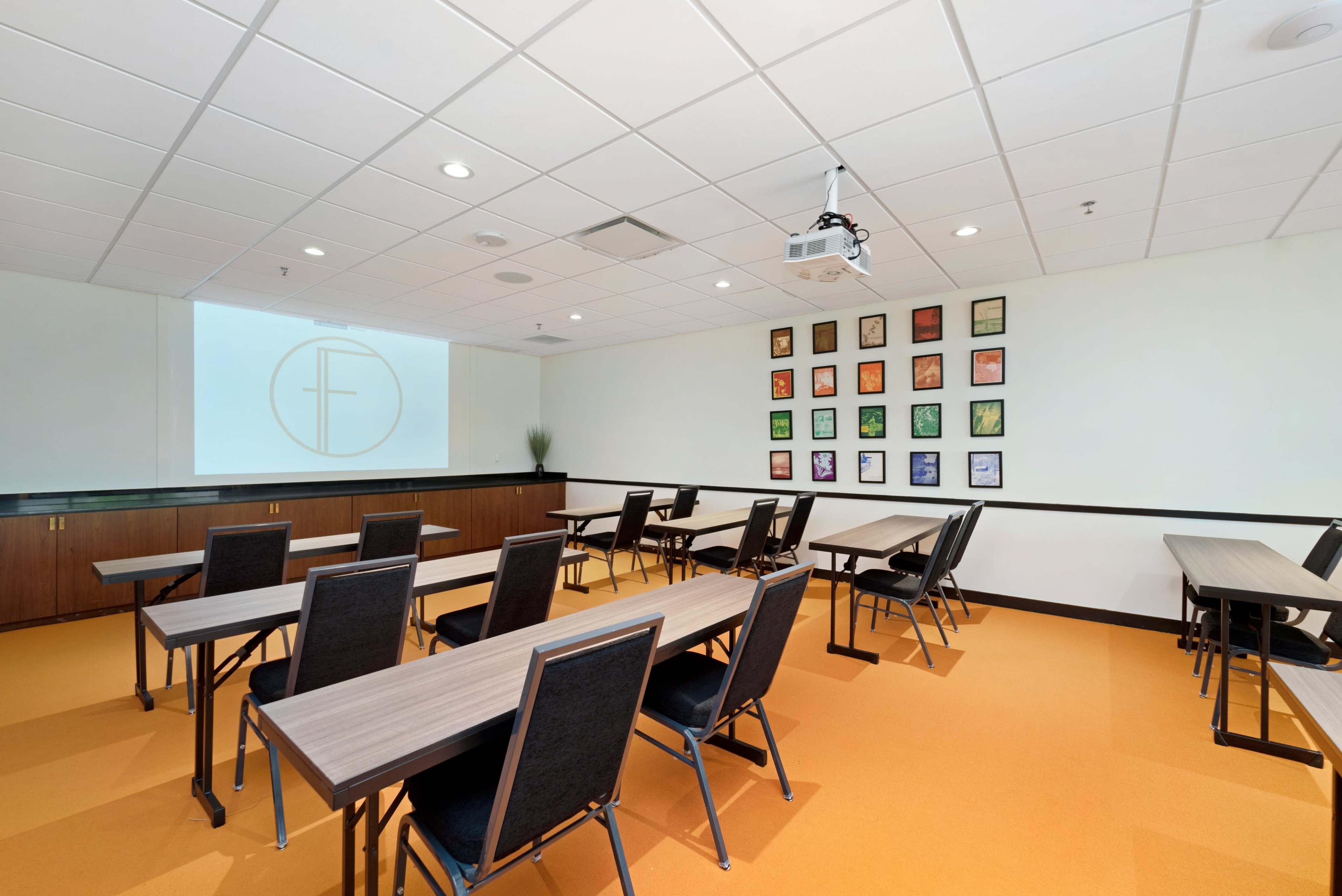 A seminar room with several rows of tables and chairs arranged facing a screen, along with colorful framed artwork on the wall.
