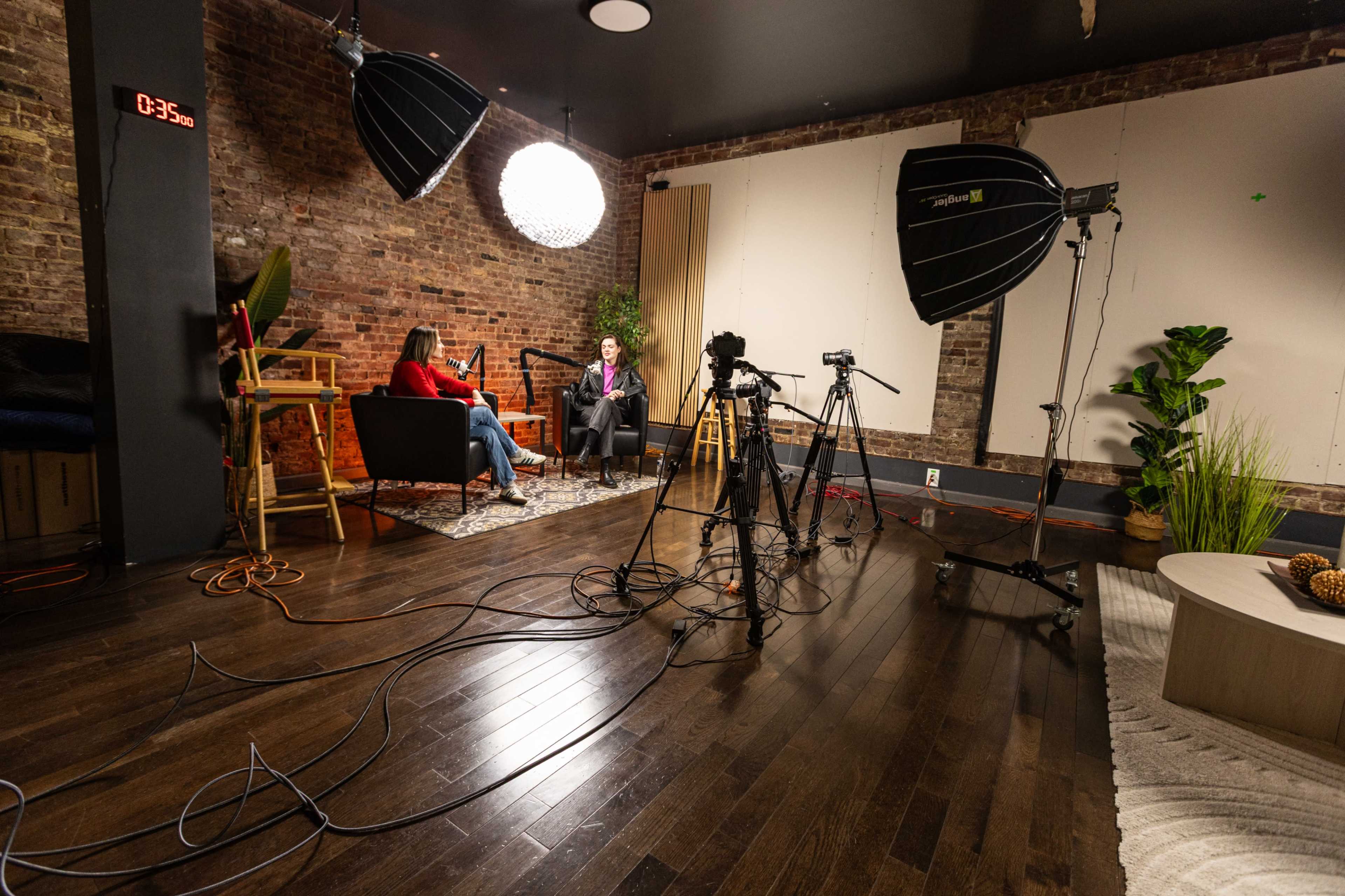 The image shows a studio setup with two women seated in chairs, surrounded by professional lighting equipment and cameras on tripods, with a brick wall in the background.