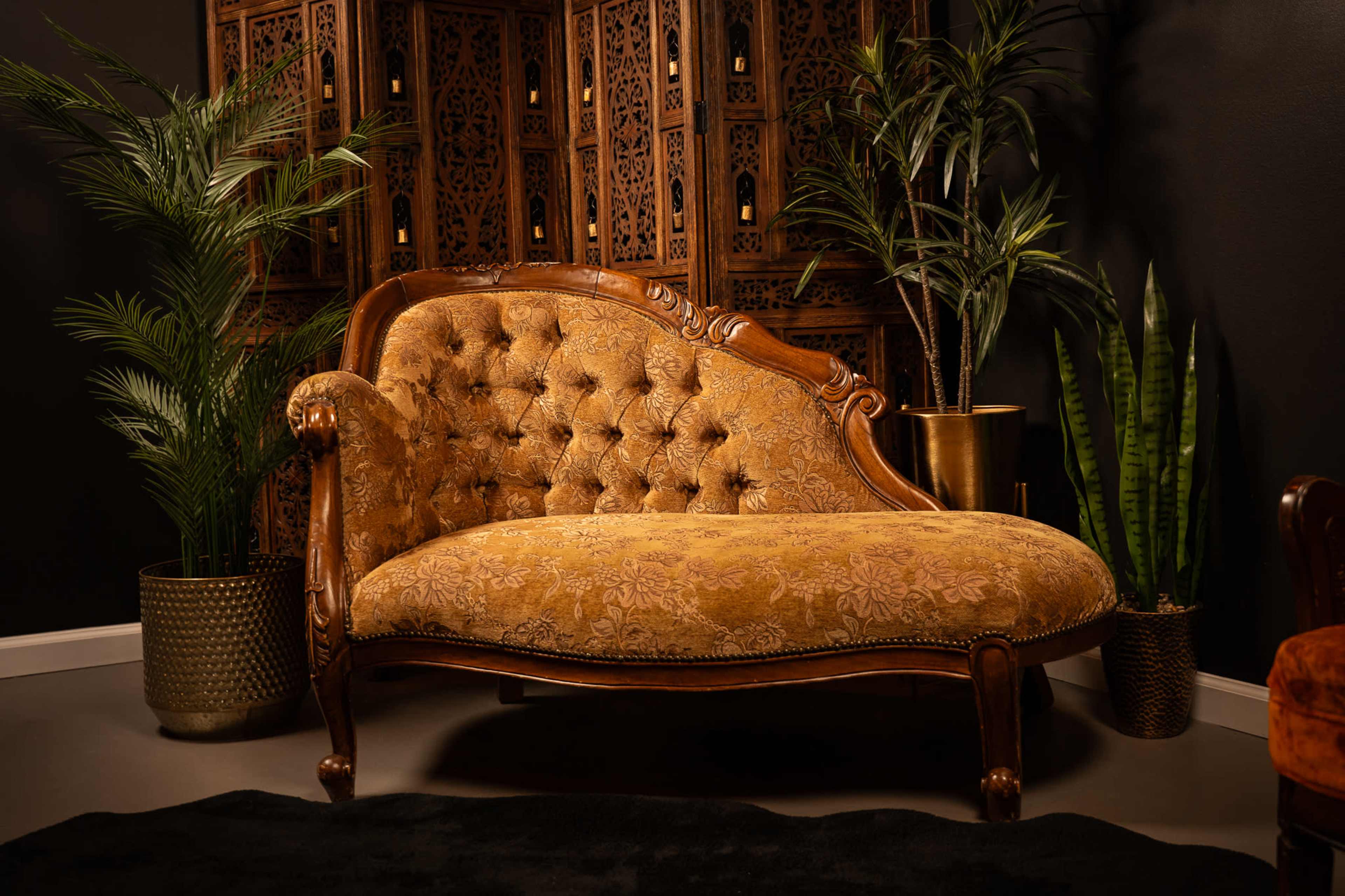 The image features an ornate Victorian-style chaise lounge with tufted upholstery, positioned against a dark wall and surrounded by decorative plants and an intricately designed wooden screen.