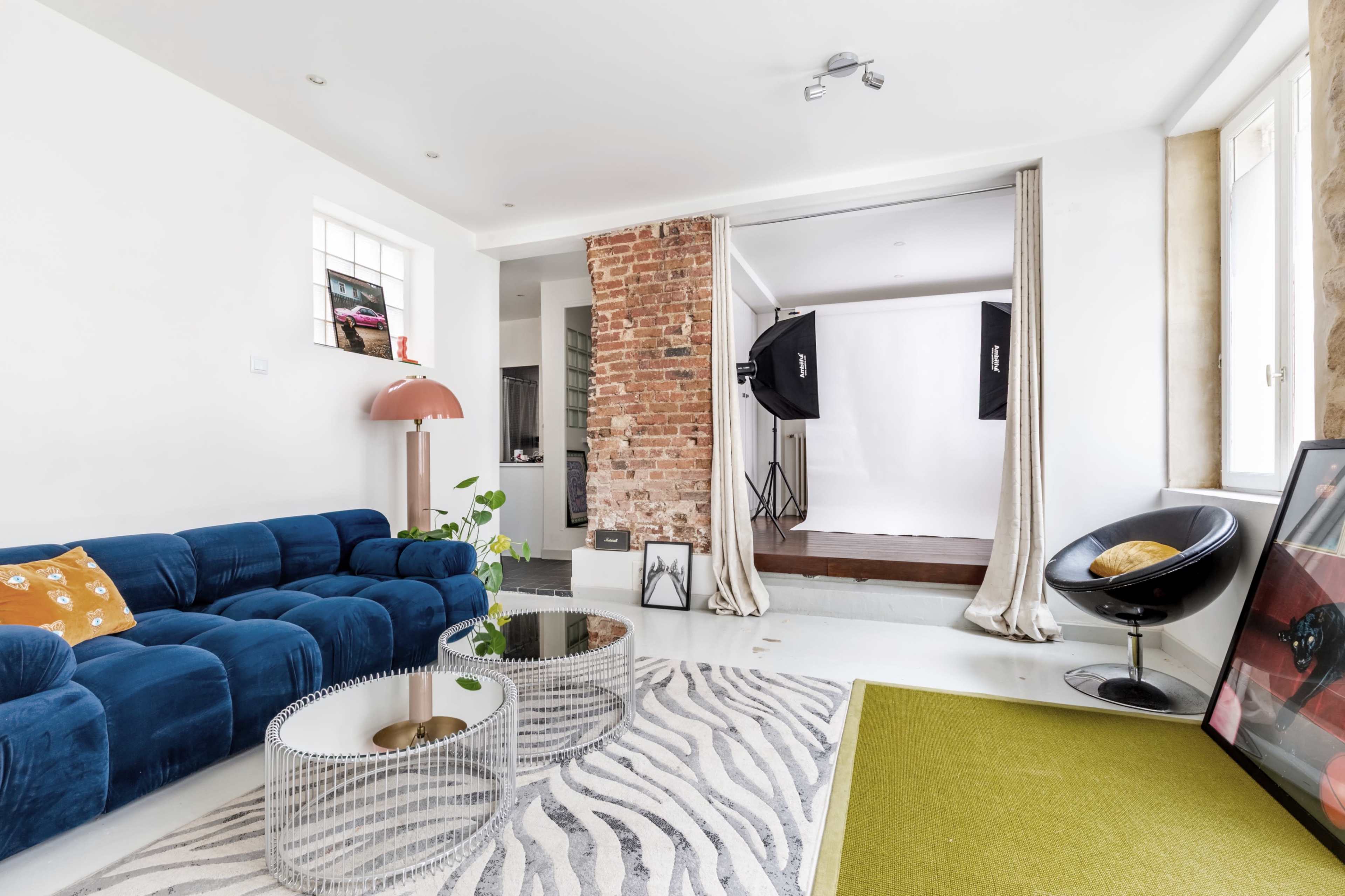 The scene features a modern living room with a blue velvet sofa, a round glass coffee table, a patterned rug, and a partially visible photography studio in the background.