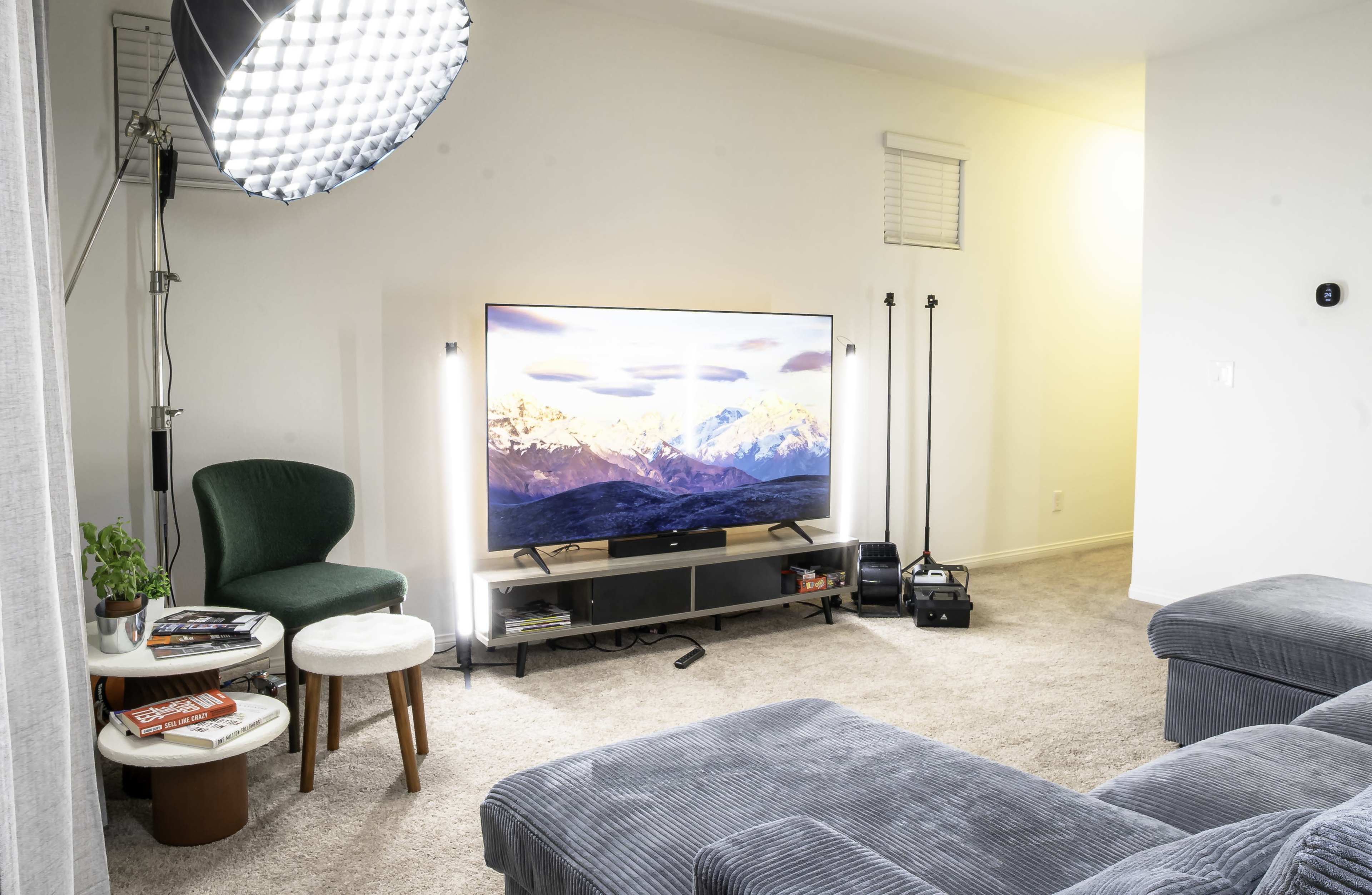 The image shows a cozy living room with a television mounted on a low cabinet, a green chair, a small side table with books and a plant, and bright lighting from a floor lamp.