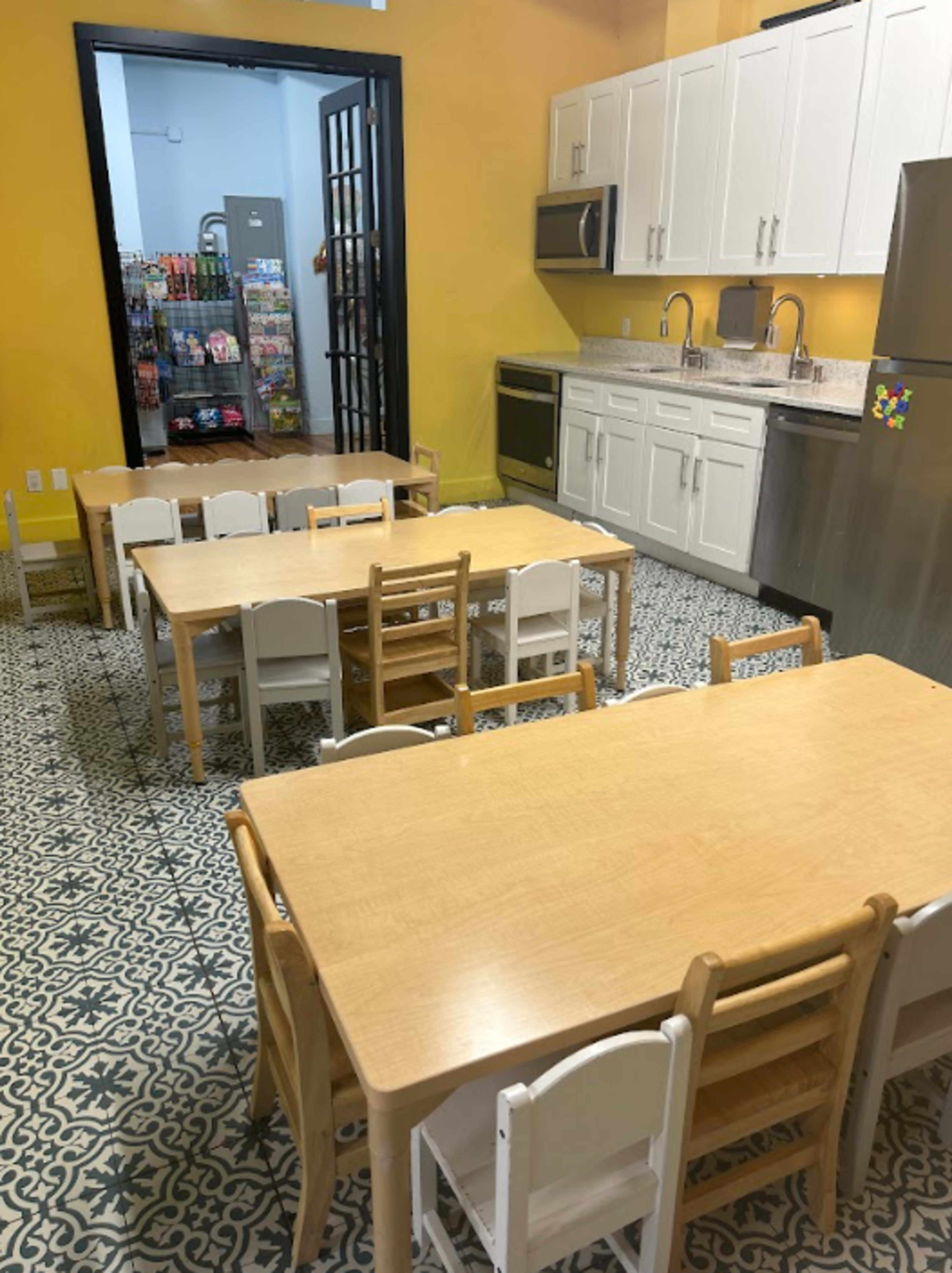 The image shows a brightly colored room with a kitchen area, featuring wooden tables and chairs arranged neatly on a patterned tiled floor.