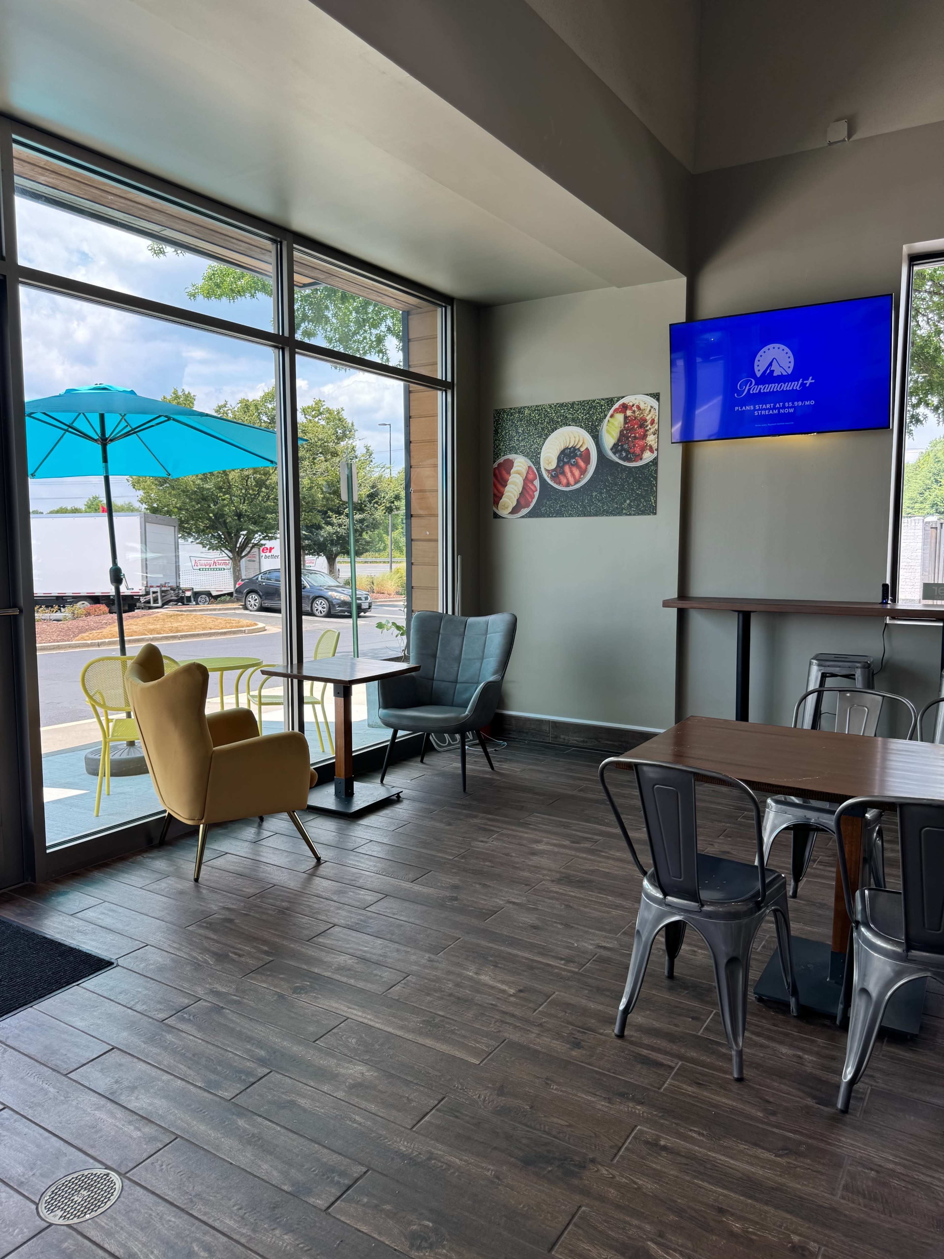 A modern cafe interior with large windows, a yellow umbrella table outside, and various seating arrangements including yellow and gray chairs.