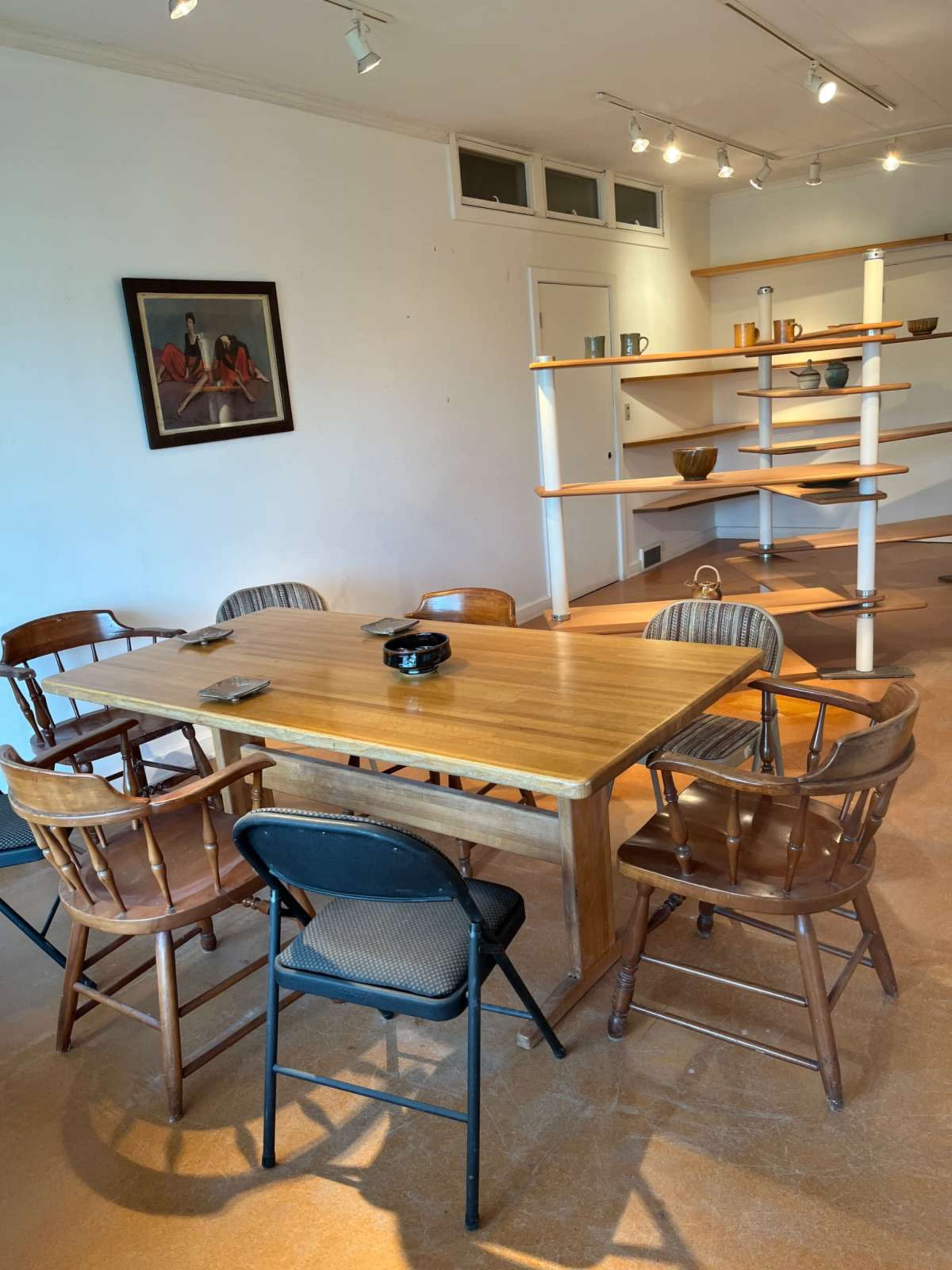 A wooden dining table surrounded by several chairs, with a painting on the wall and a shelf displaying items in the background.