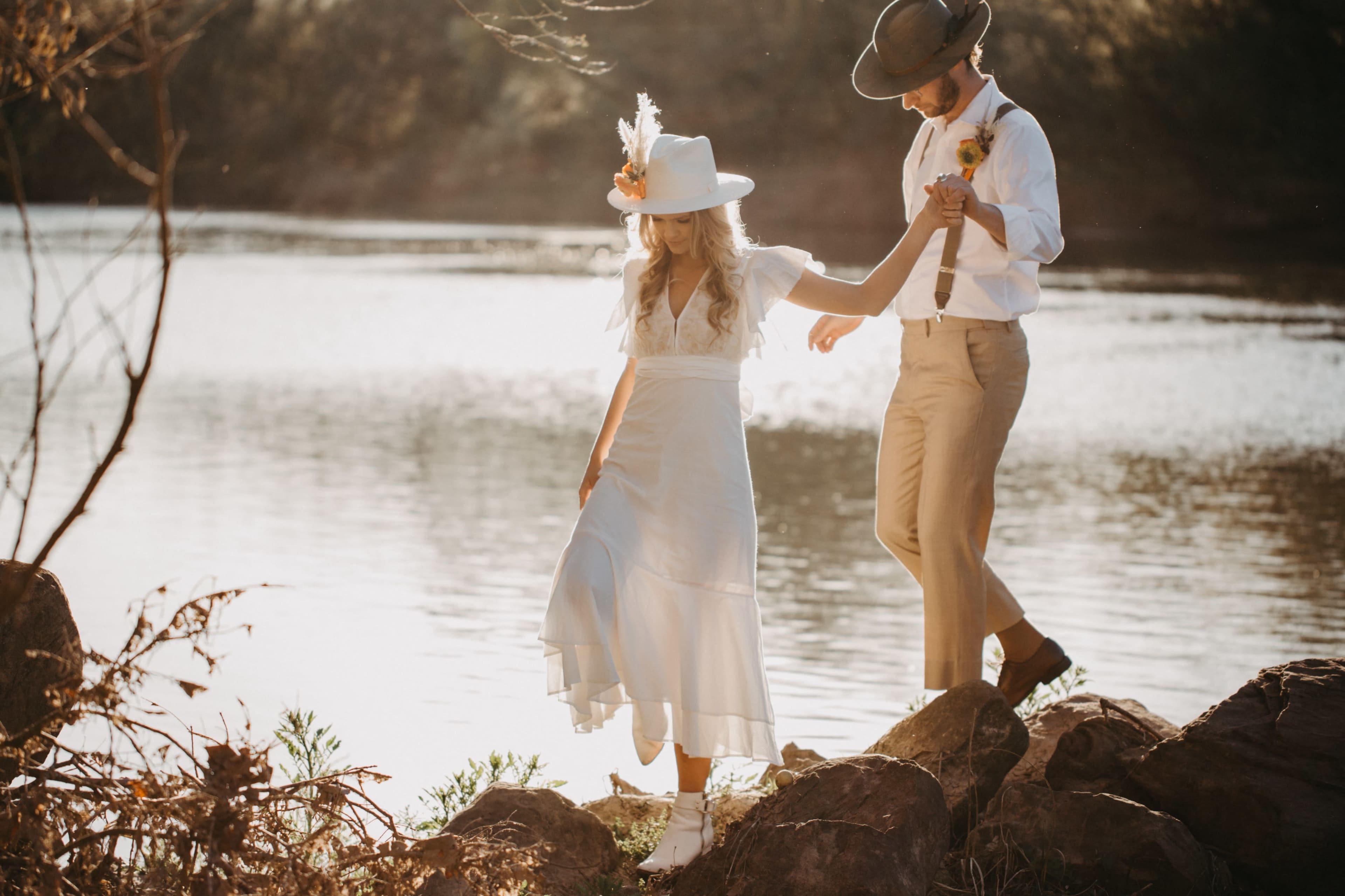 A couple walks hand in hand along the rocky shore of a river, with the sun setting in the background.