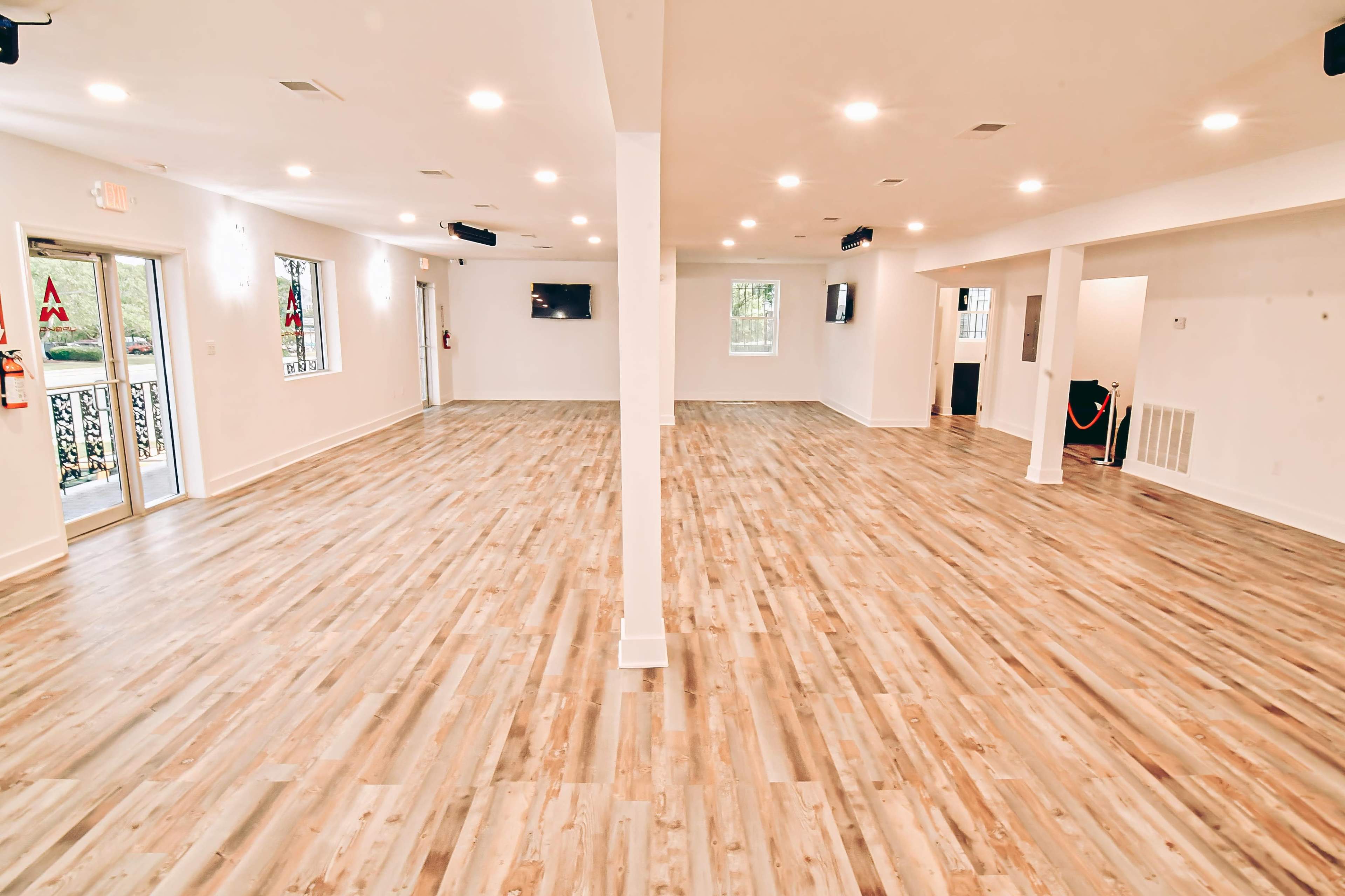 The image shows a spacious, well-lit room with light-colored wood flooring, divided by a central support column and featuring large windows.