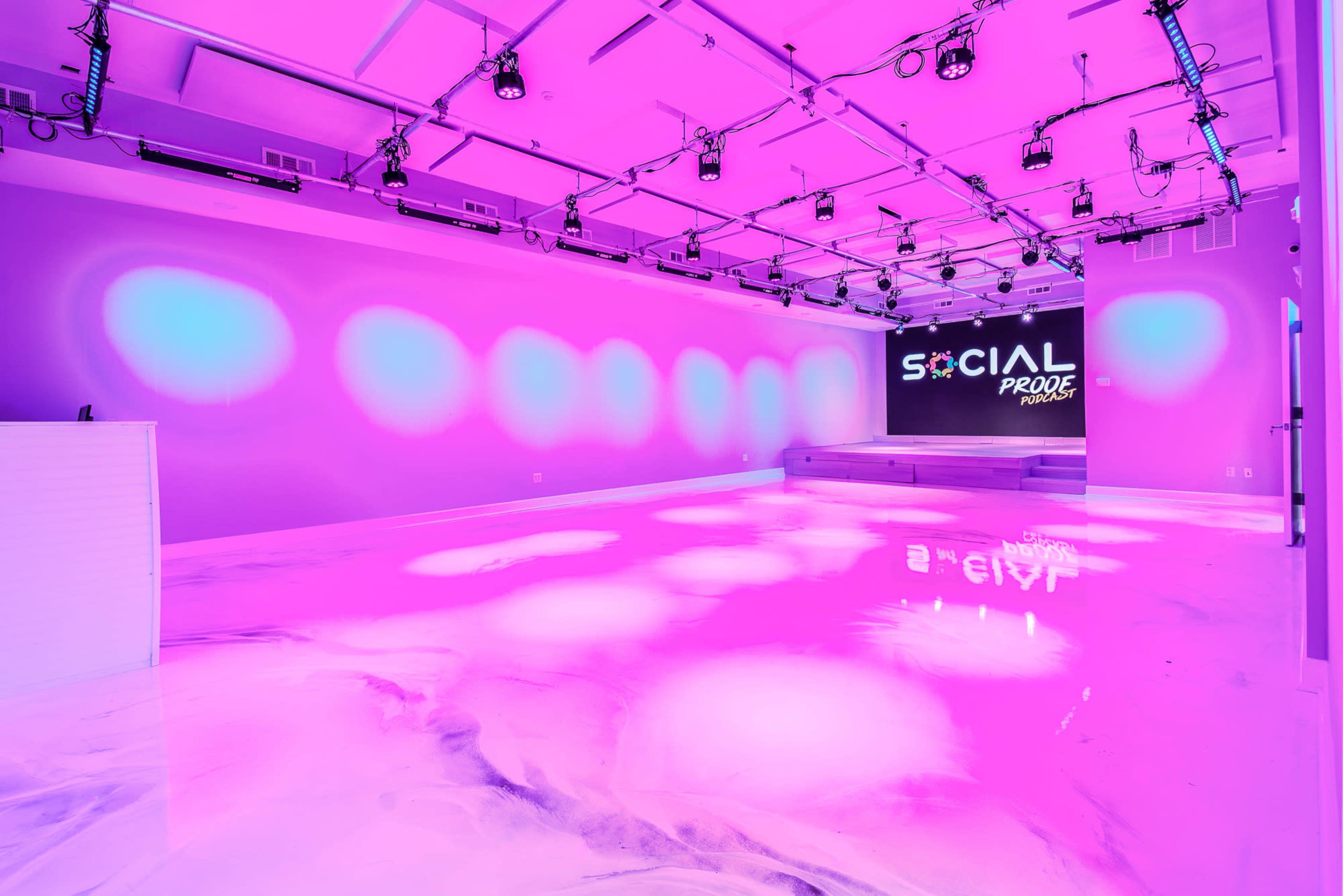 A brightly lit studio space features a glossy floor and pink-hued walls, with a large screen displaying the text "SOCIAL PRODE" on one side.