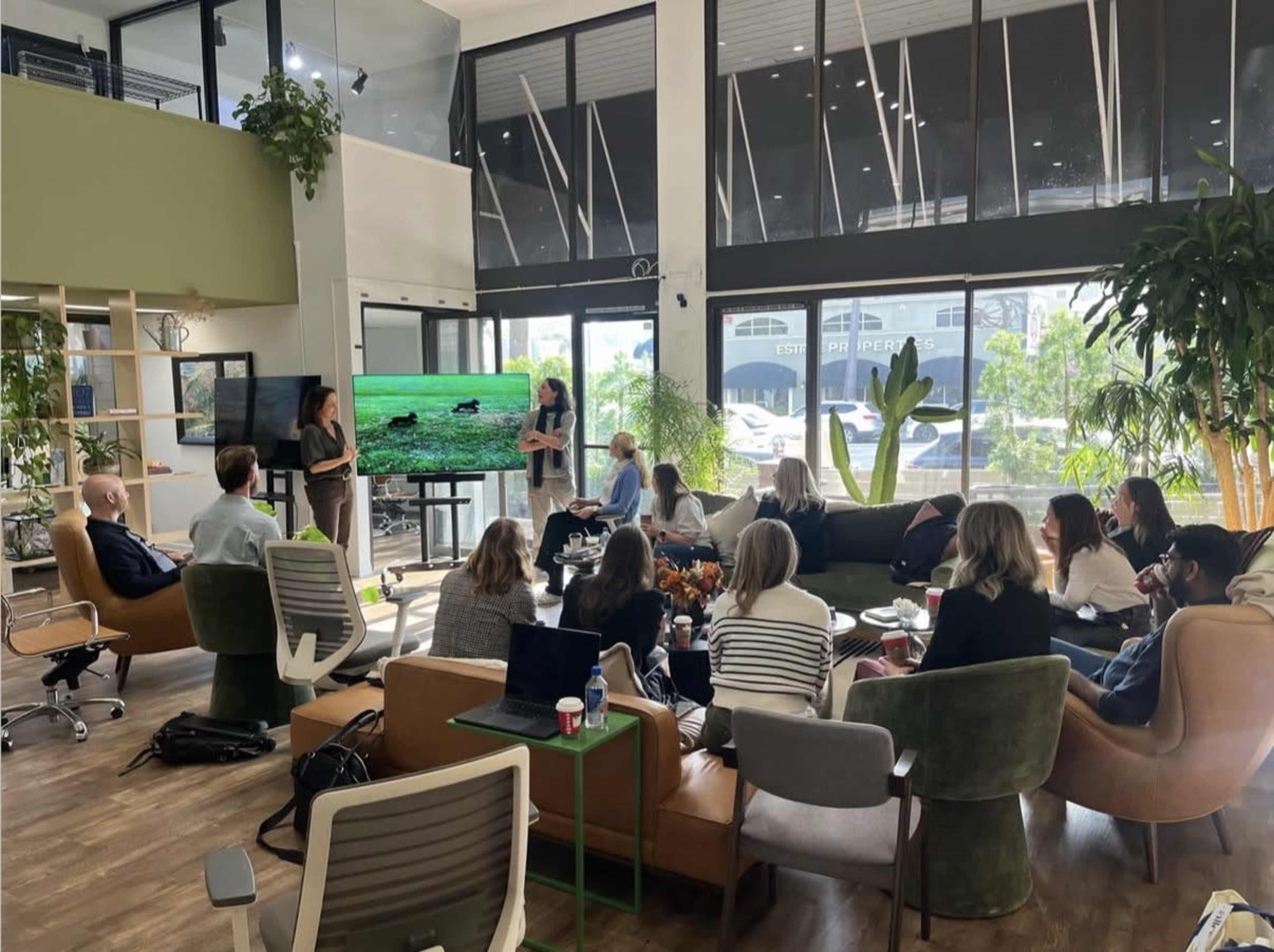 A group of people is sitting and standing in a modern office space, engaged in a presentation displayed on a screen.