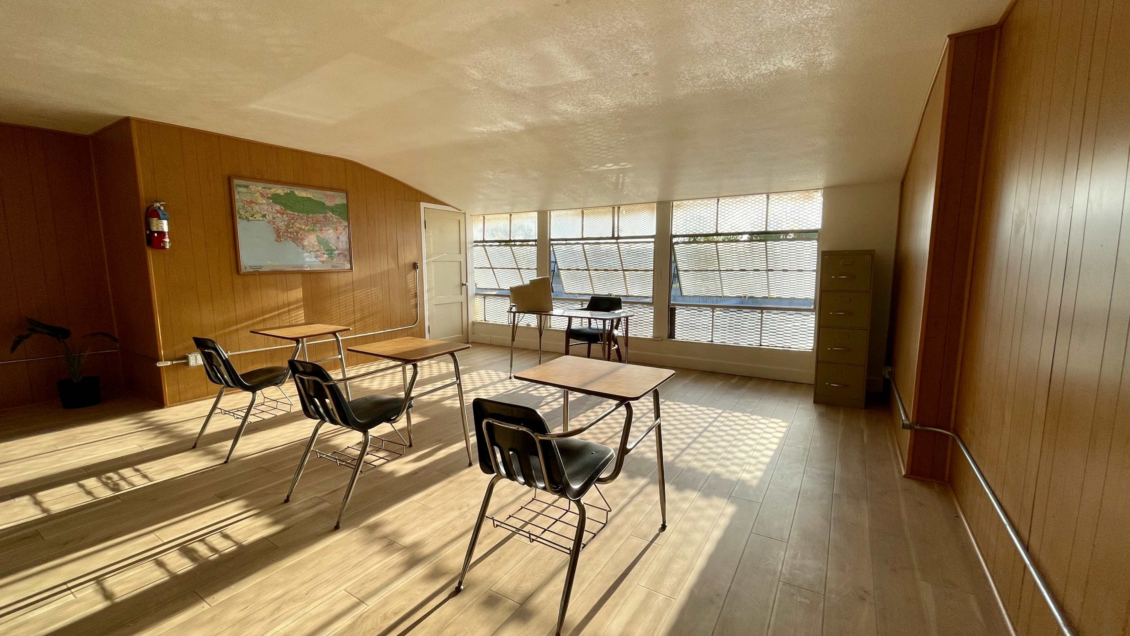 A classroom features four empty desks arranged in a sunlit room with wooden walls and large windows.