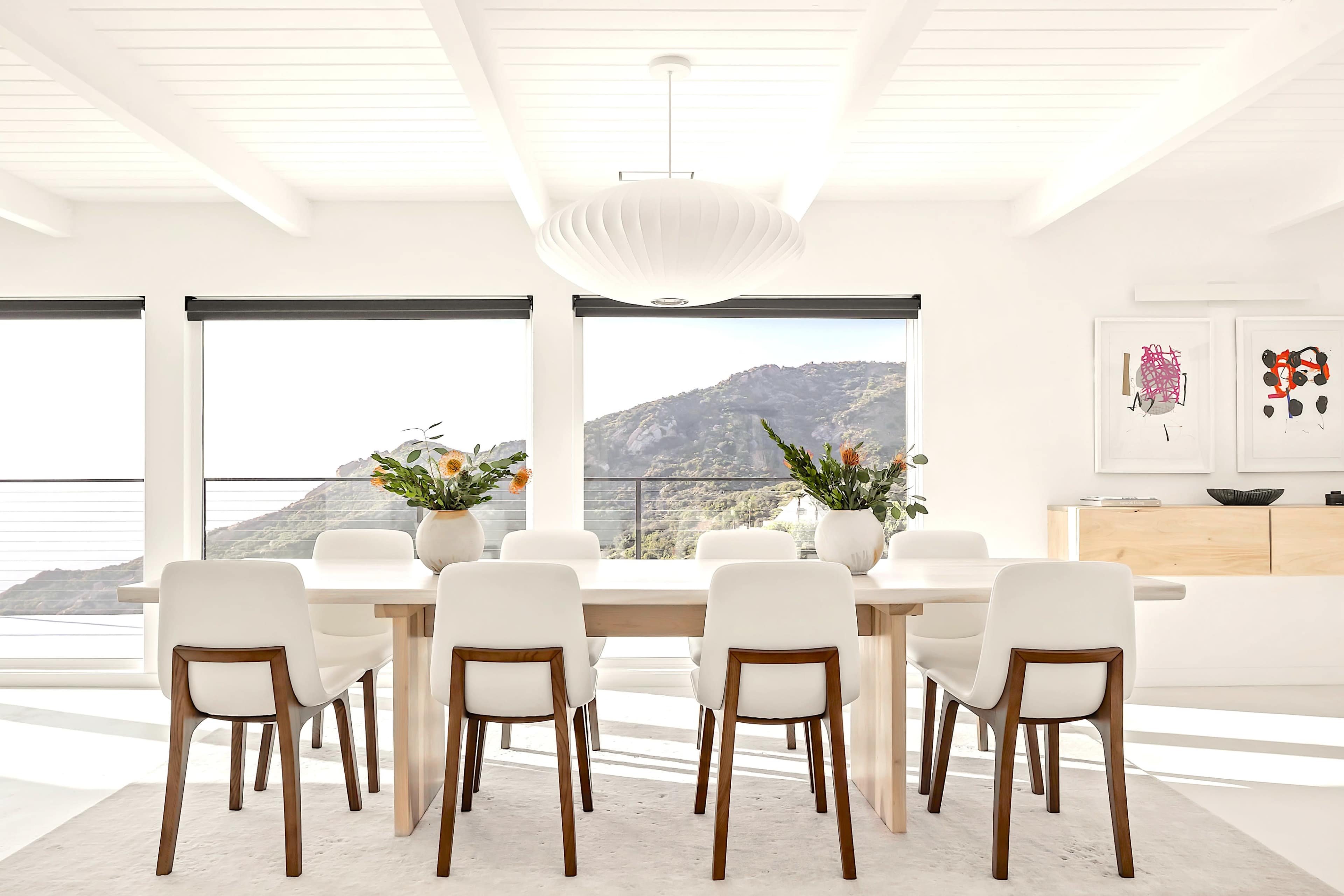 A spacious dining room features a long wooden table surrounded by white chairs, large windows revealing a mountain view, and modern decor.