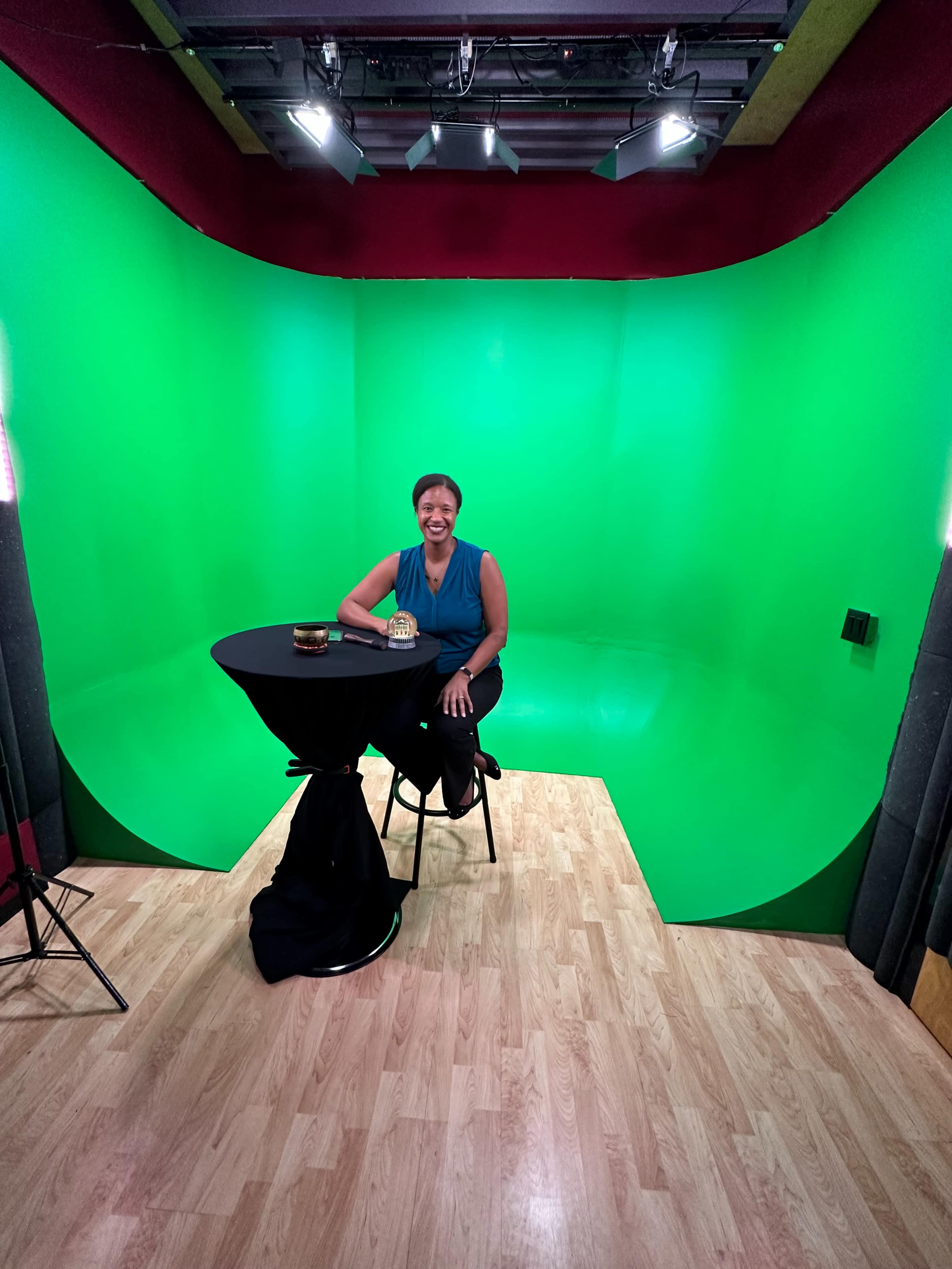 A person sits at a black table in a studio with a green backdrop and wooden flooring.