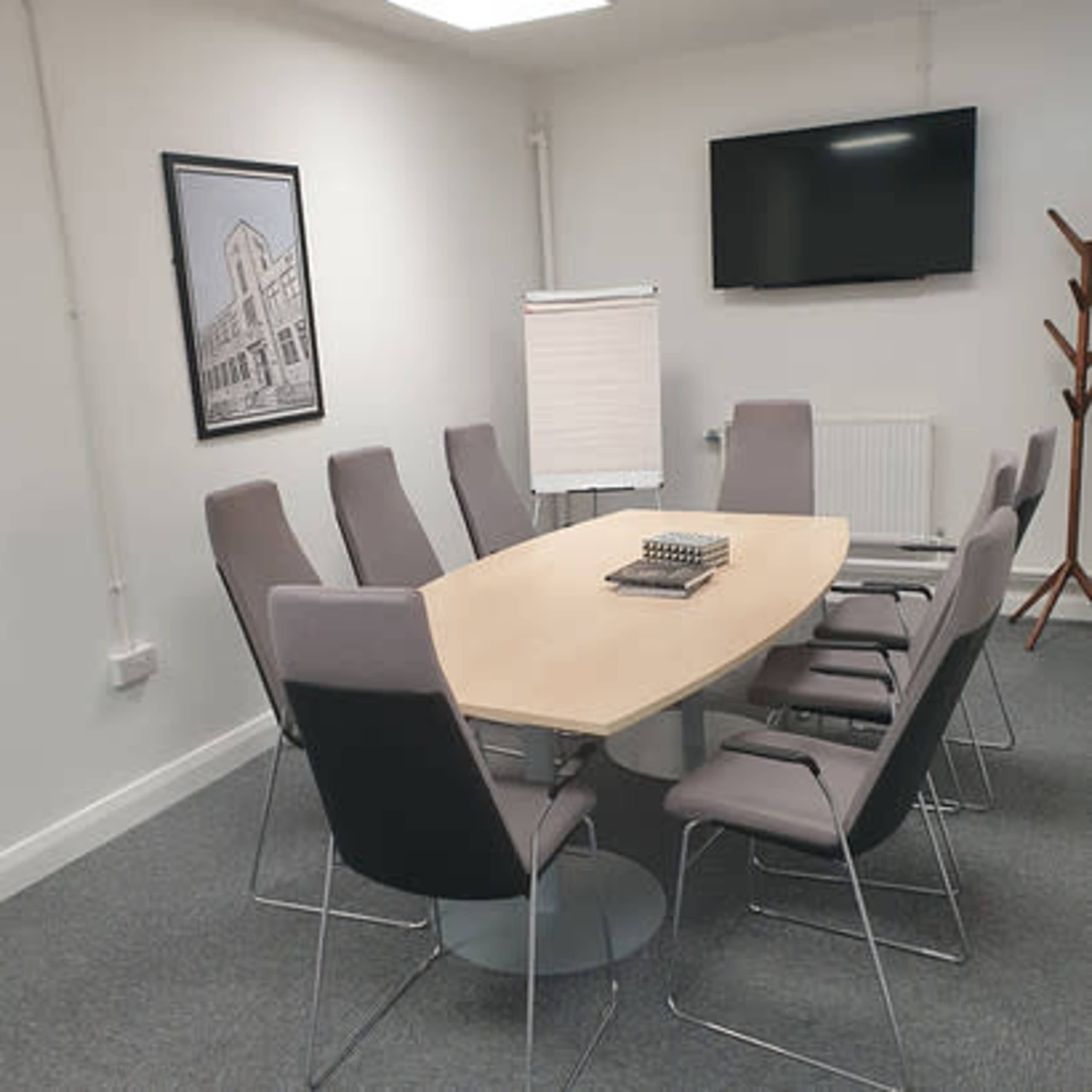 A modern conference room features a large oval table surrounded by eight chairs, with a flip chart, television, and a wall-mounted artwork.