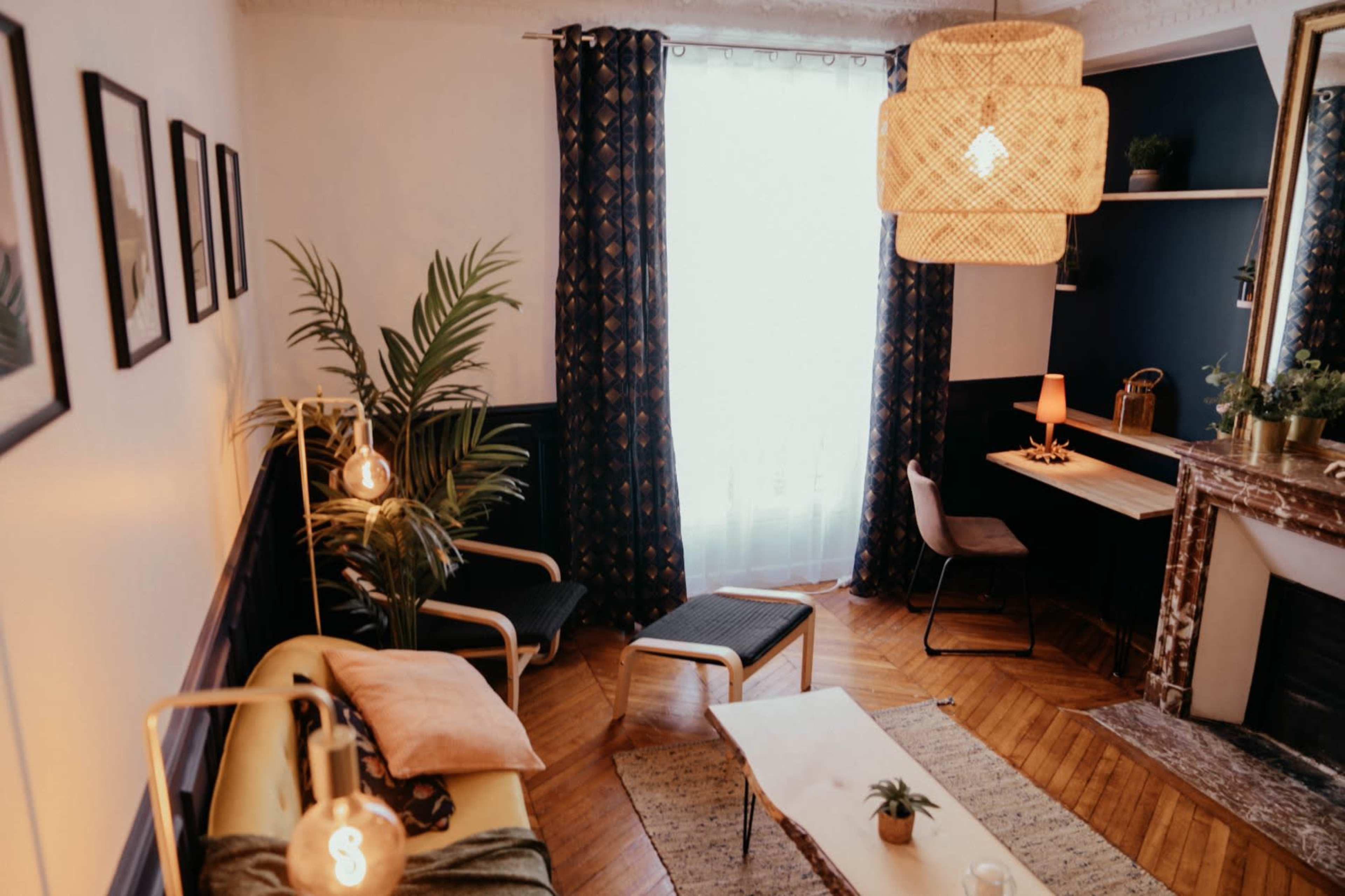 The image shows a cozy living room with a wooden floor, a large window draped with curtains, and various plants, furniture, and decor elements.