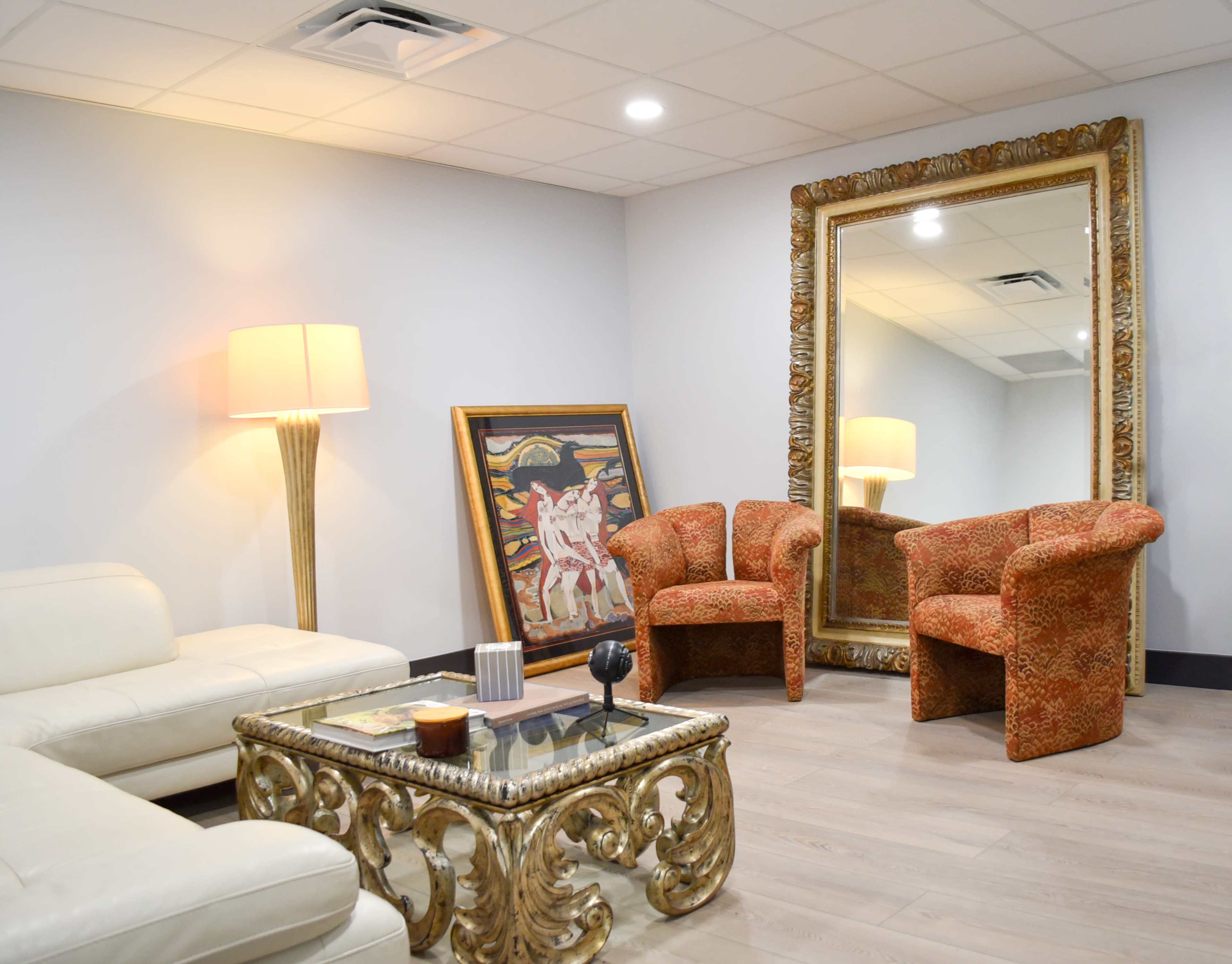 The image shows a modern lounge area featuring a white leather sofa, two patterned chairs, a decorative mirror, and a glass coffee table.