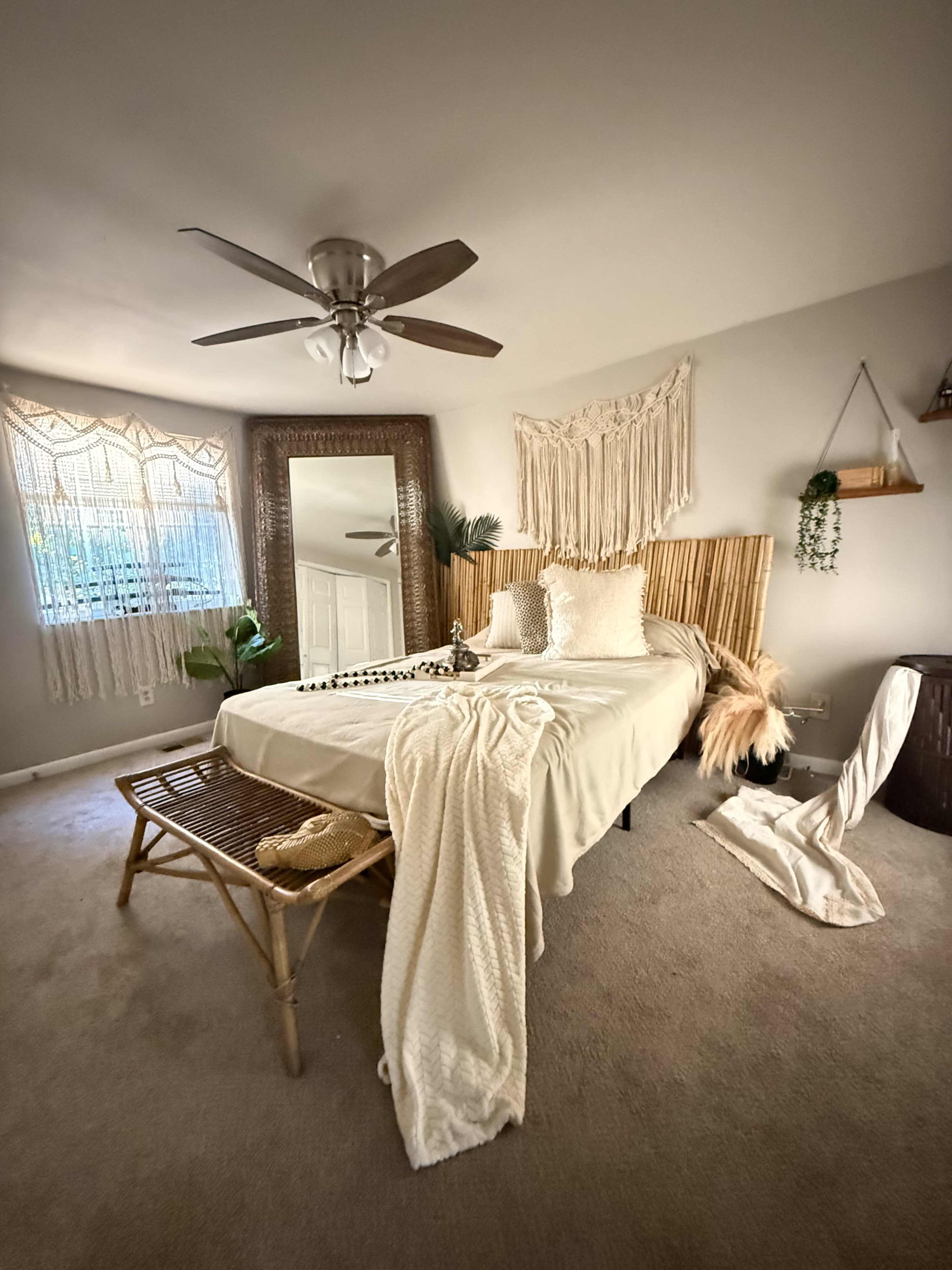 The image shows a cozy bedroom featuring a bed with a woven headboard, a large mirror, and decorative plants, all set against light-colored walls and soft carpeting.