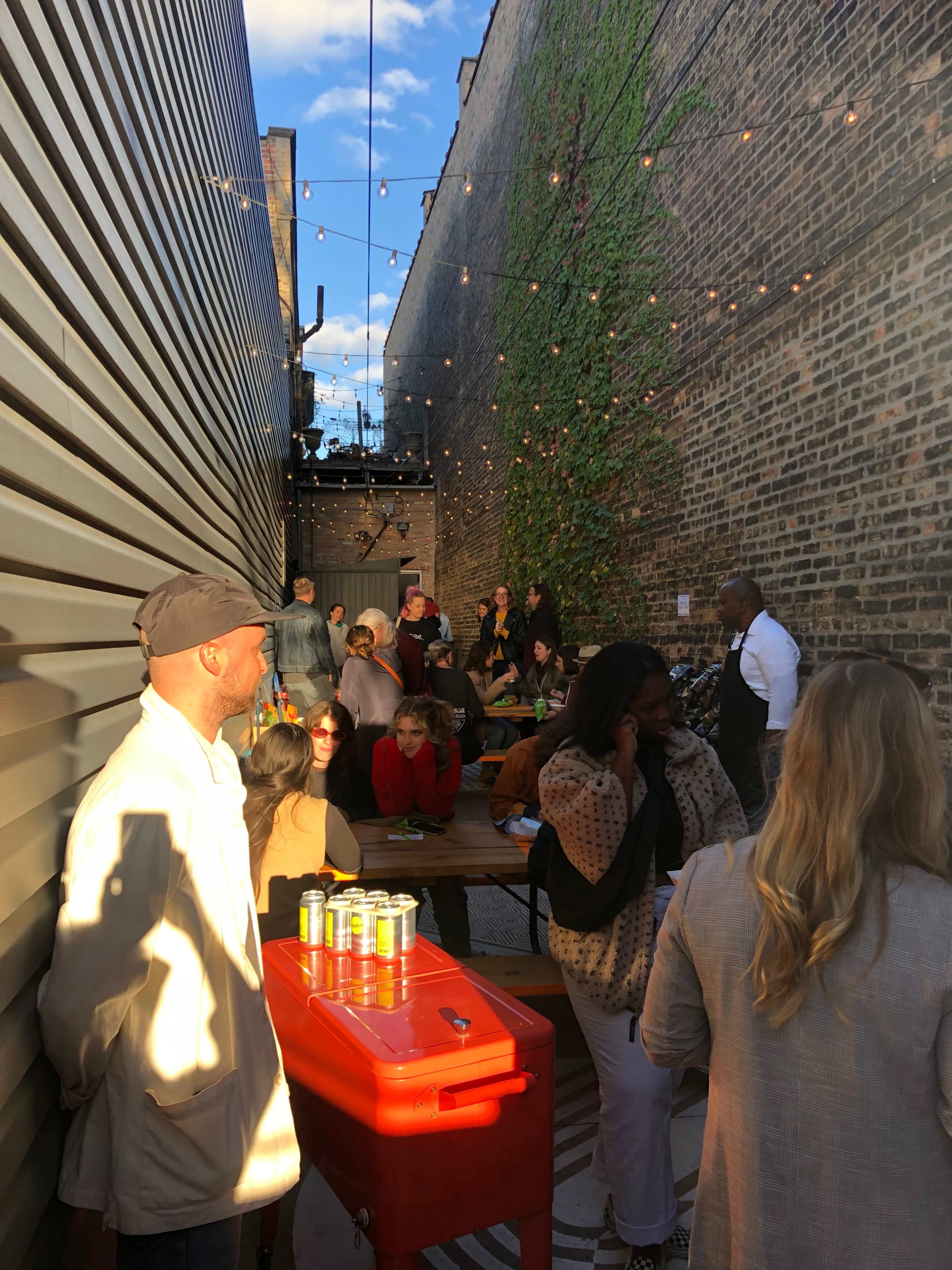 A gathering of people in an outdoor alleyway adorned with string lights, where some are seated at tables and others are mingling near a cooler.