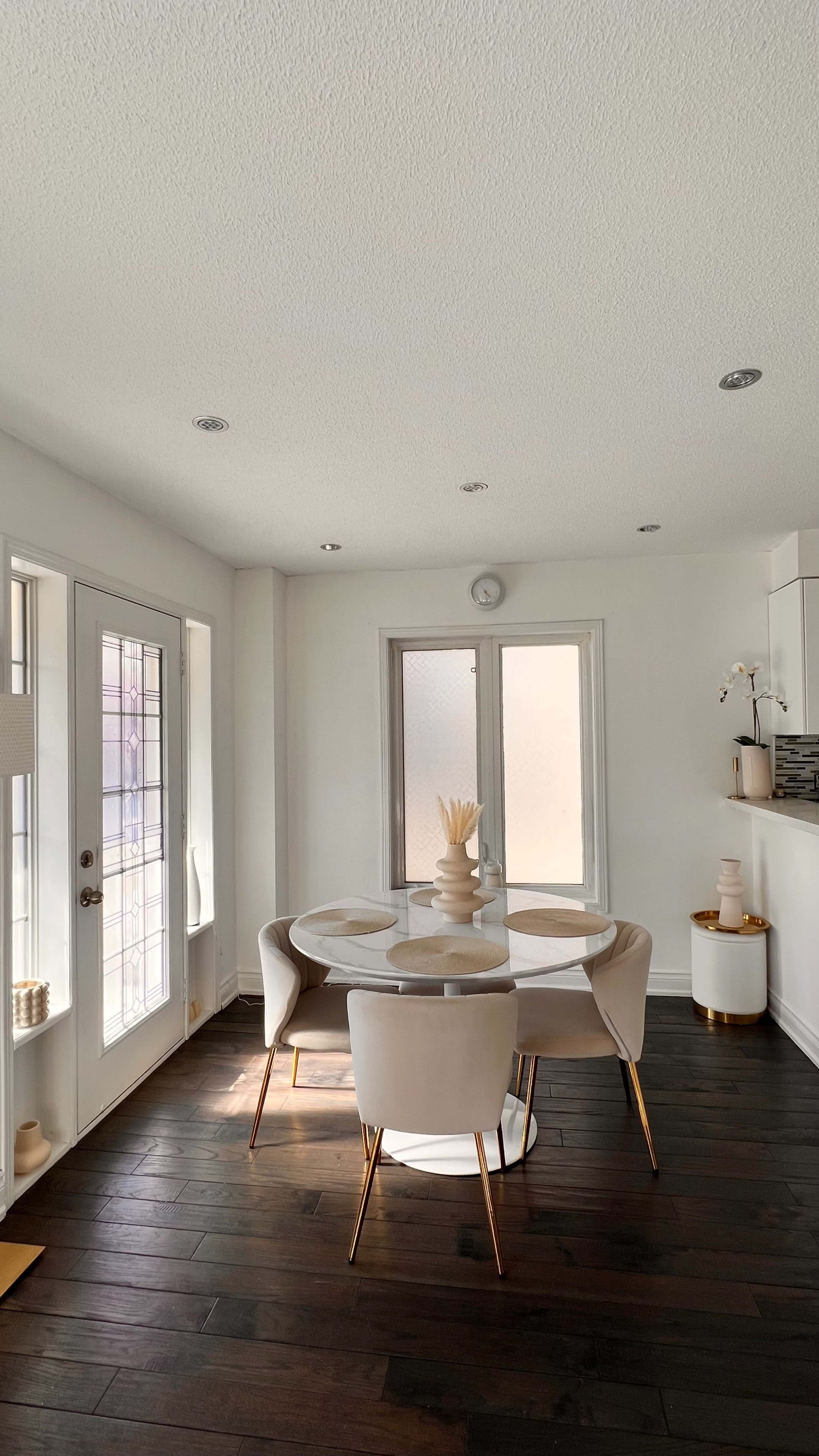 A modern dining area features a circular table with four white chairs, situated near large windows and wooden flooring.