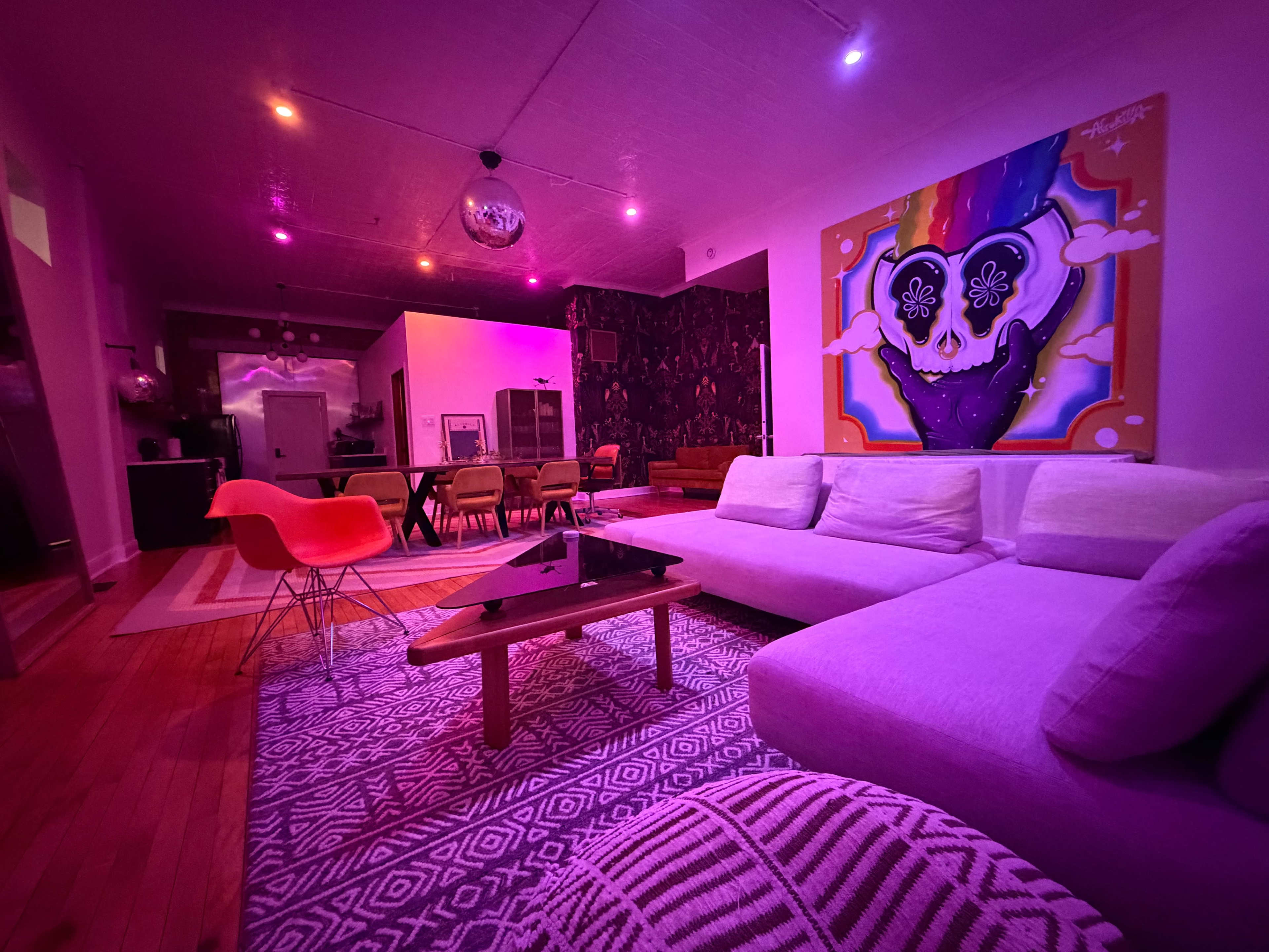 The image depicts a vibrant, modern living space with purple lighting, a large white sofa, a patterned rug, and a colorful wall mural featuring a heart design.