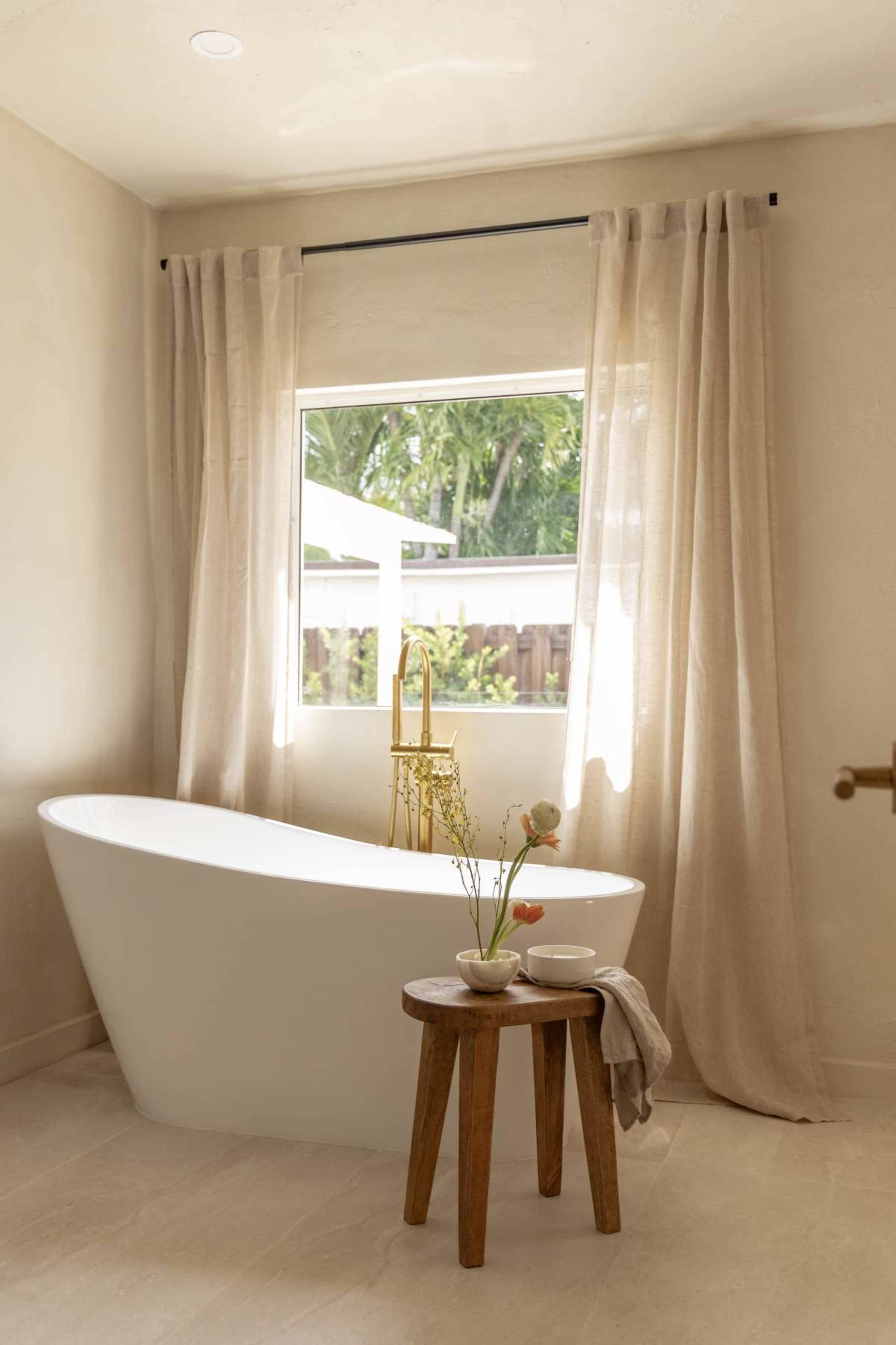A bright bathroom features a freestanding white bathtub, a small wooden stool with decorative items, and sheer curtains framing a window.