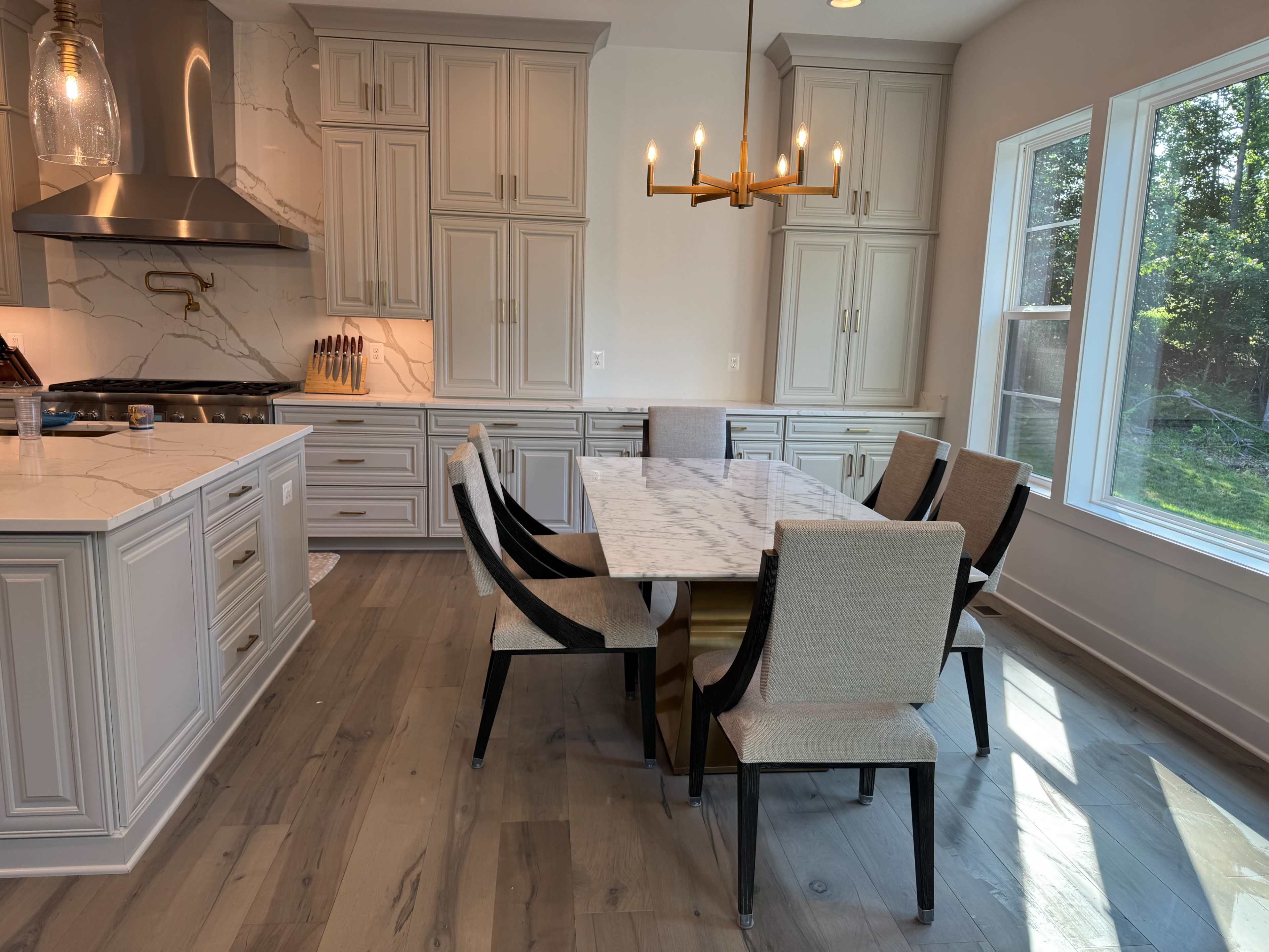 The image shows a modern kitchen with gray cabinetry, a marble dining table surrounded by six chairs, and large windows that overlook a green area.
