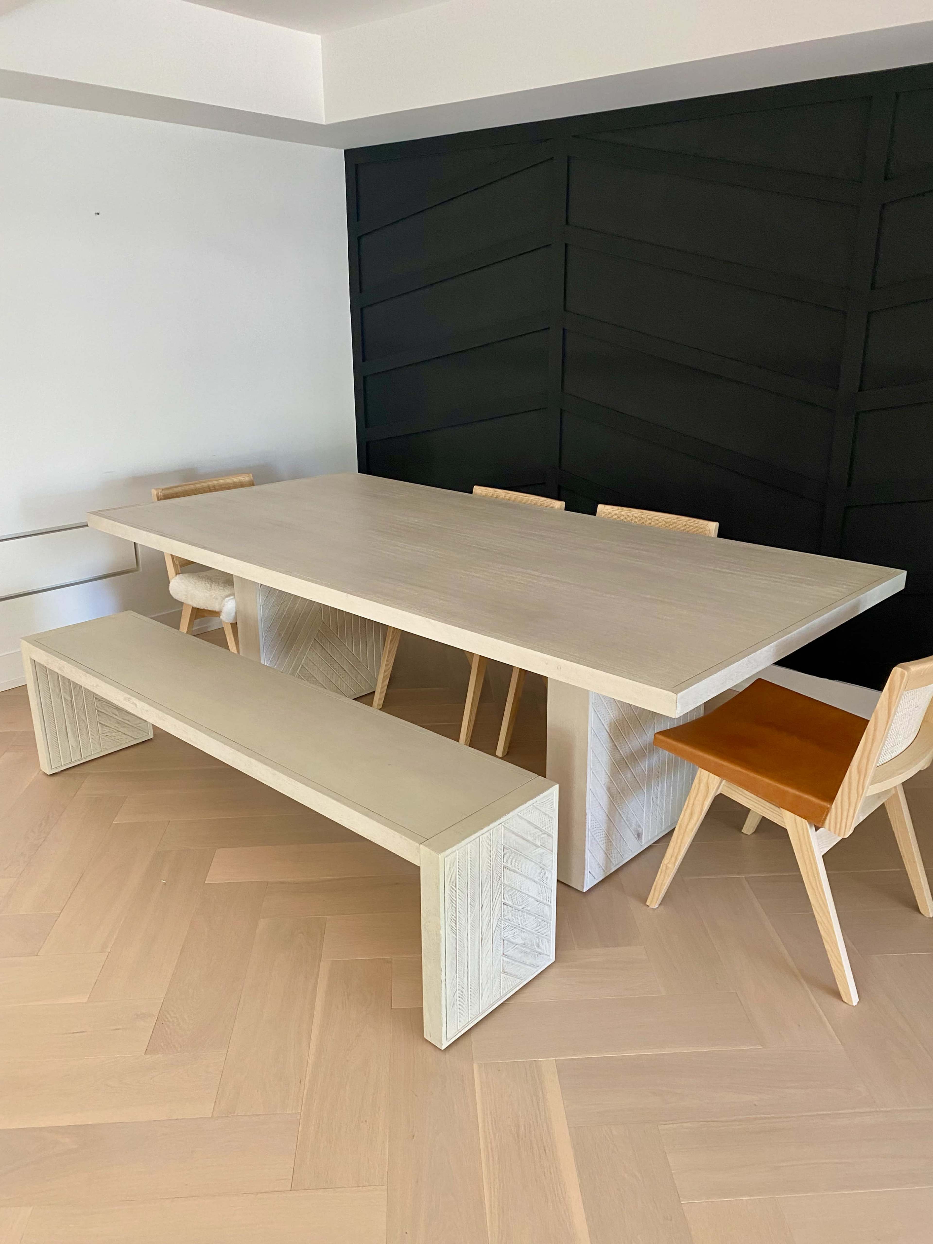 A rectangular dining table with a light wooden finish is surrounded by wooden chairs and a long bench in a modern interior with a dark accent wall.