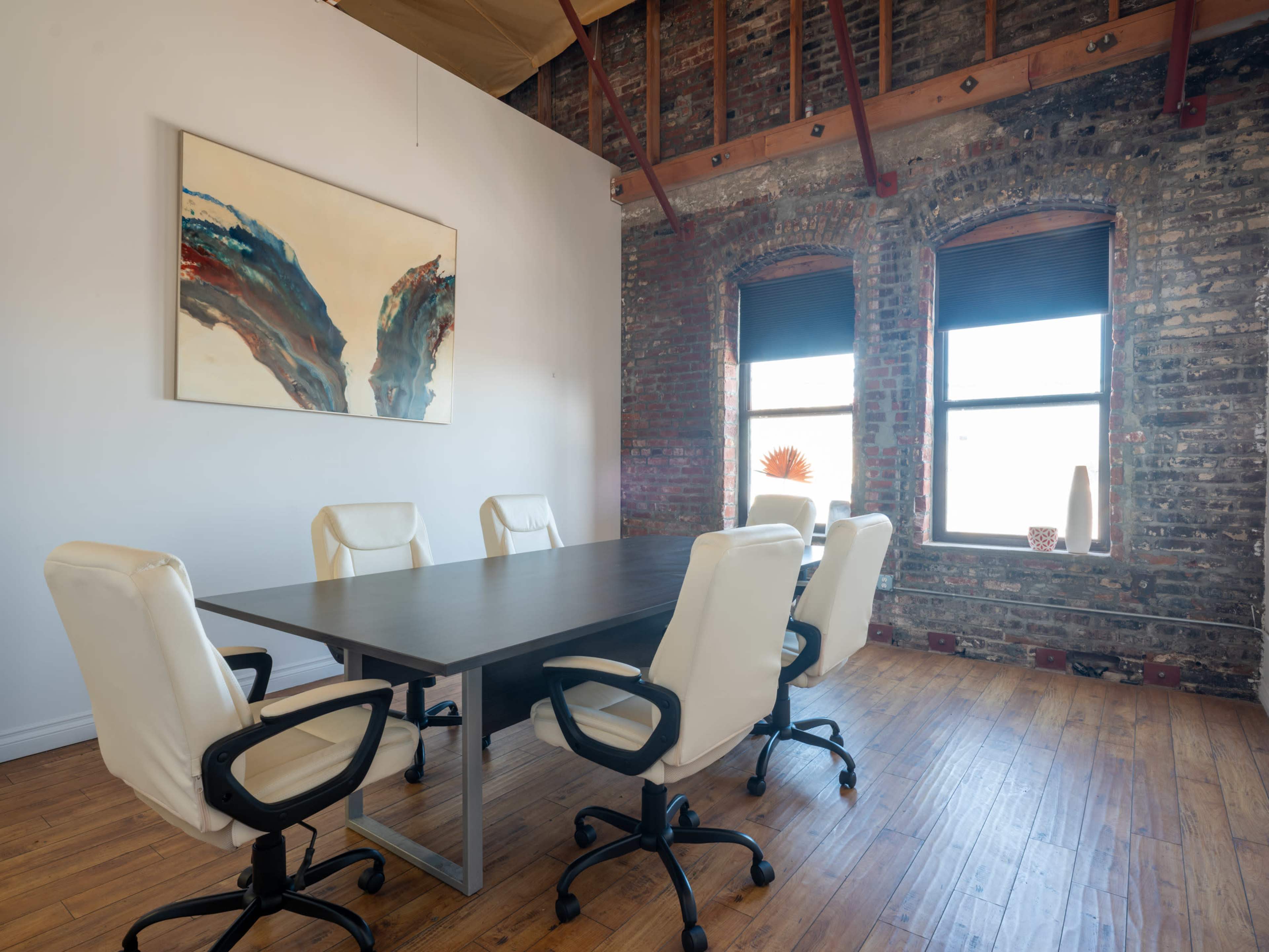 A modern conference room features a long table surrounded by white leather chairs, with exposed brick walls and large windows.
