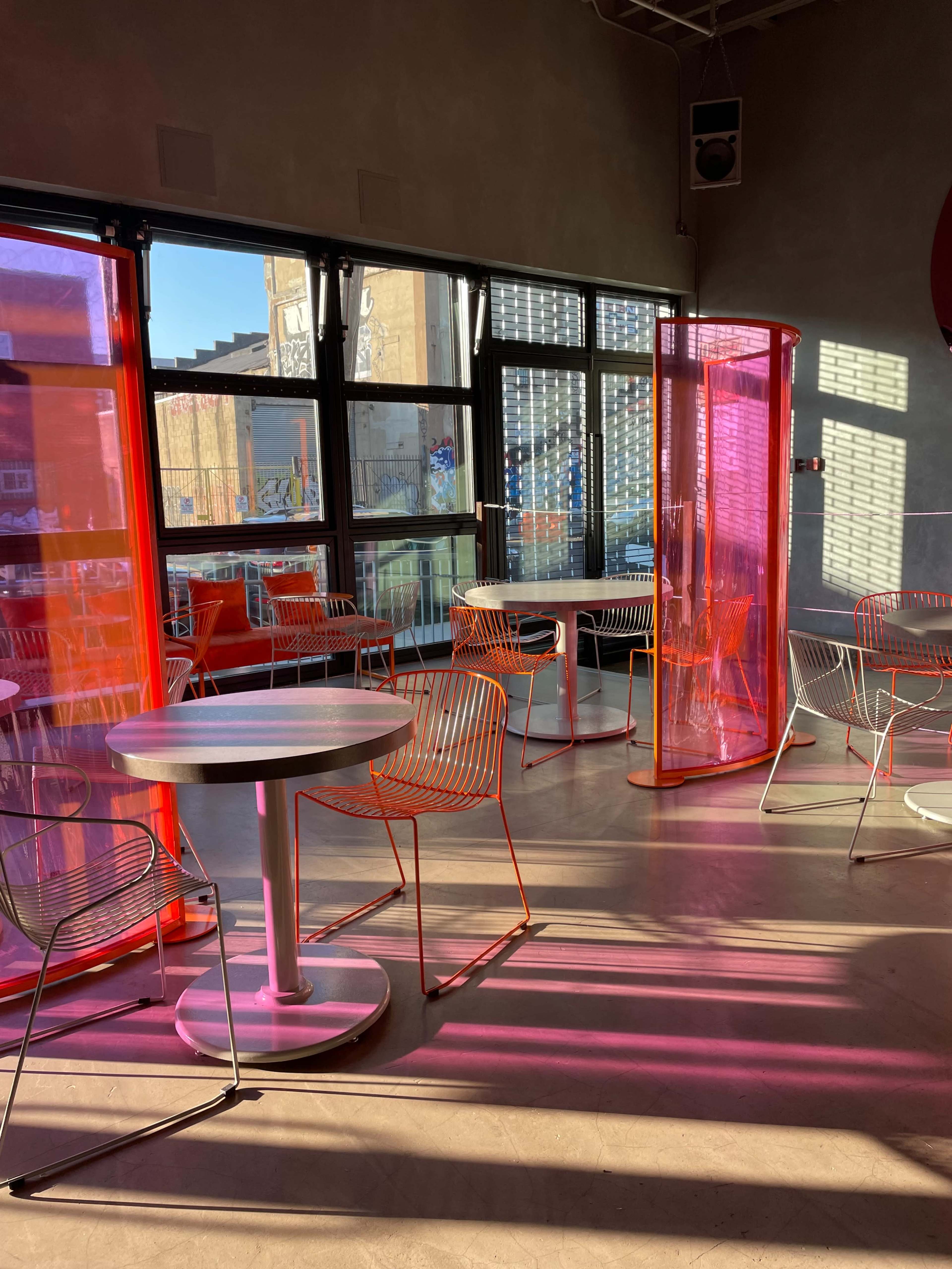 The image shows a modern café interior with colorful translucent partitions, empty tables, and sunlight casting shadows across the floor.