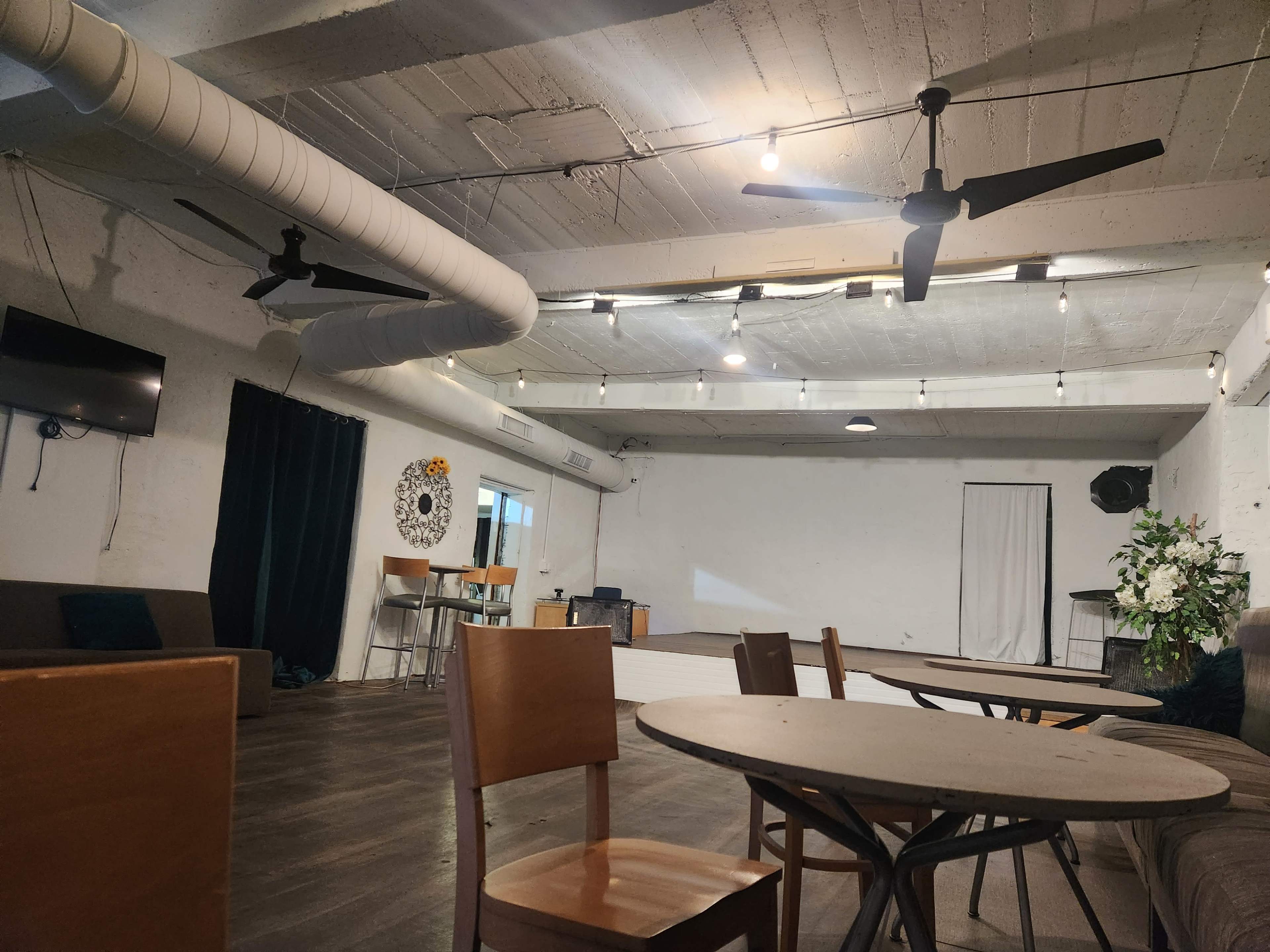 An empty venue with wooden tables and chairs, ceiling fans, and a large wall-mounted screen.