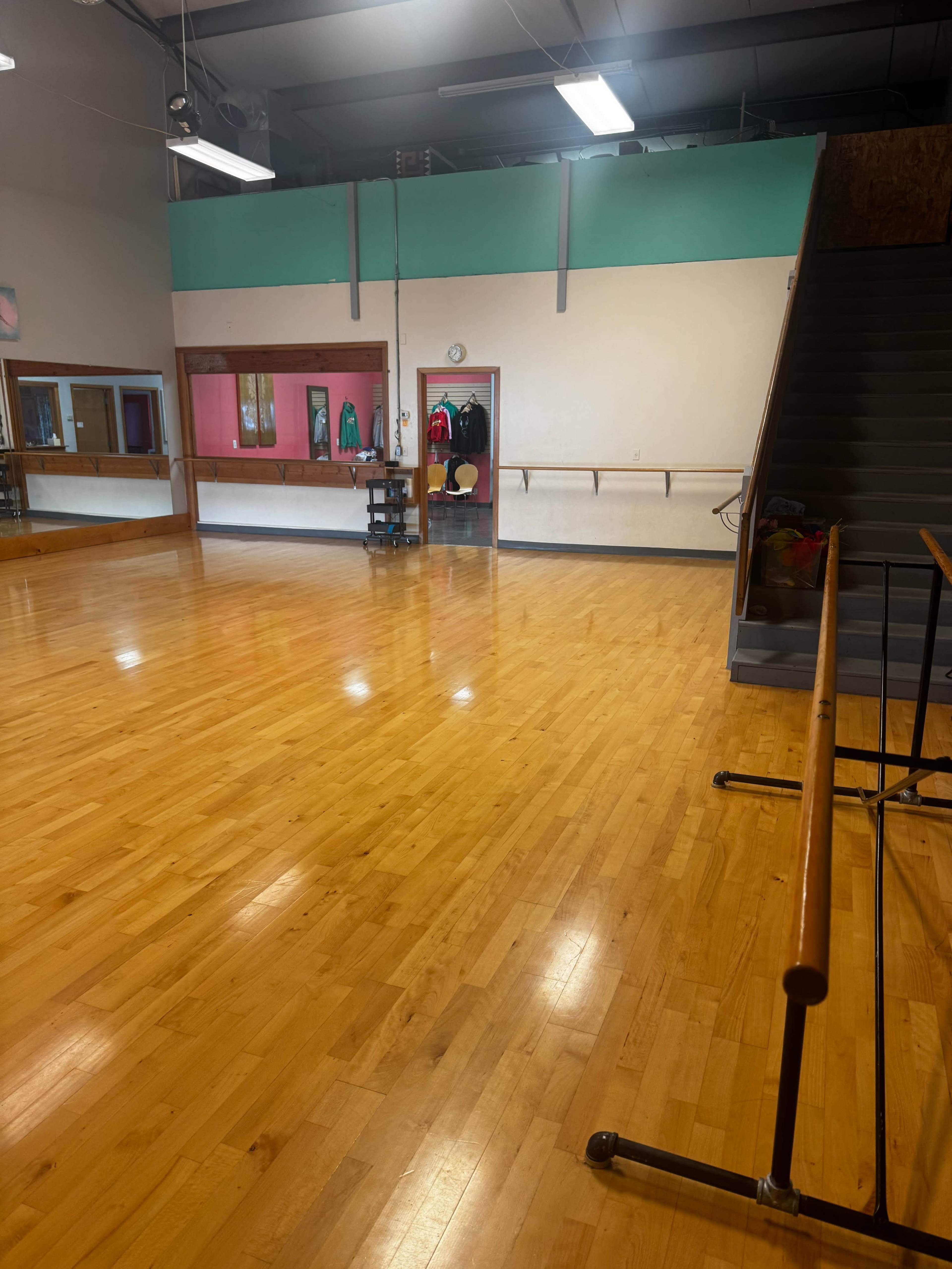 The image shows a spacious dance studio with a polished wooden floor, mirrors along the walls, and a staircase leading to an upper level.