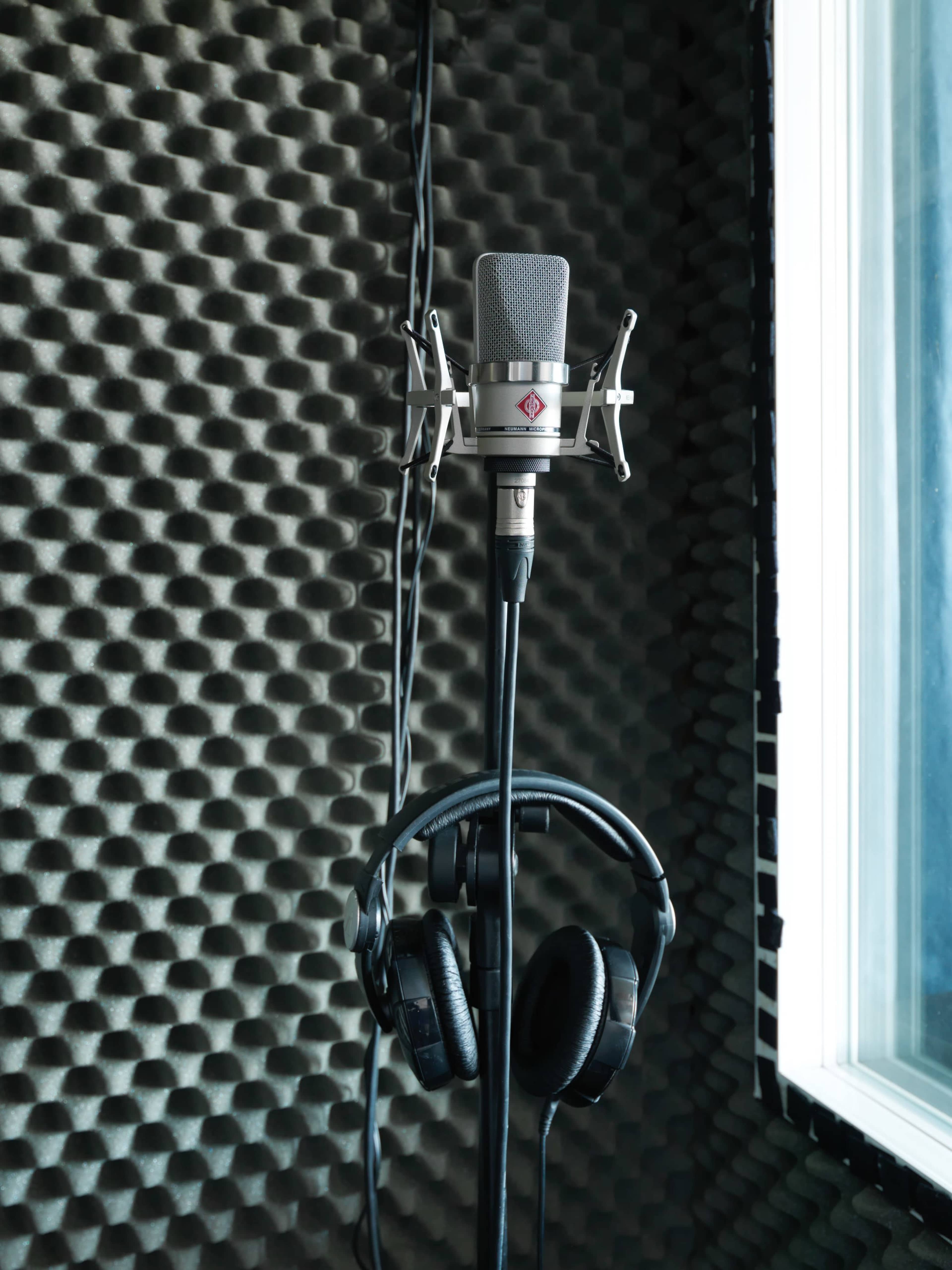 A microphone stands on a stand with headphones hanging from it, set against a textured acoustic panel backdrop near a window.