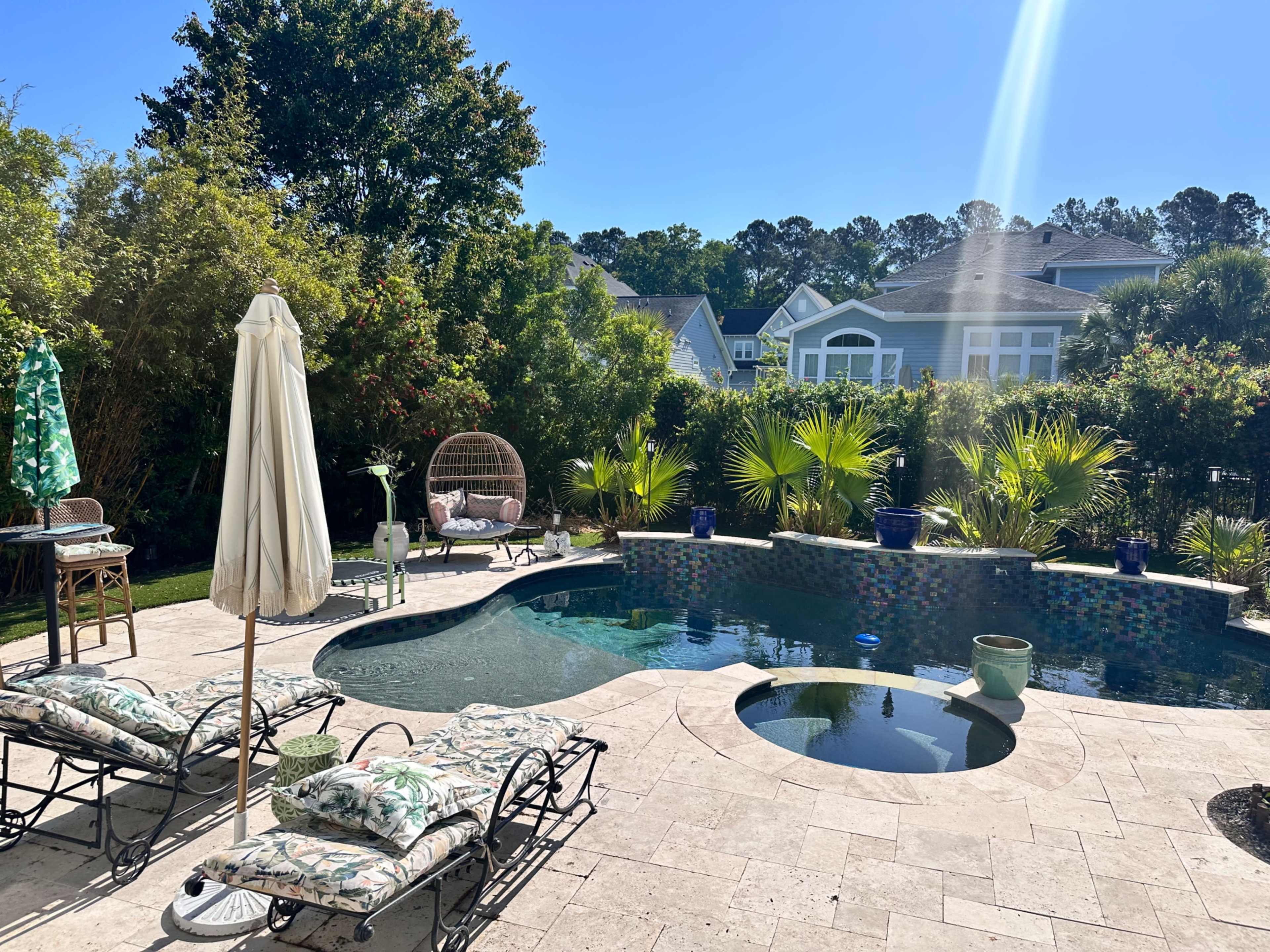 The image shows a backyard with a swimming pool, lounge chairs, a sun umbrella, and surrounding greenery.