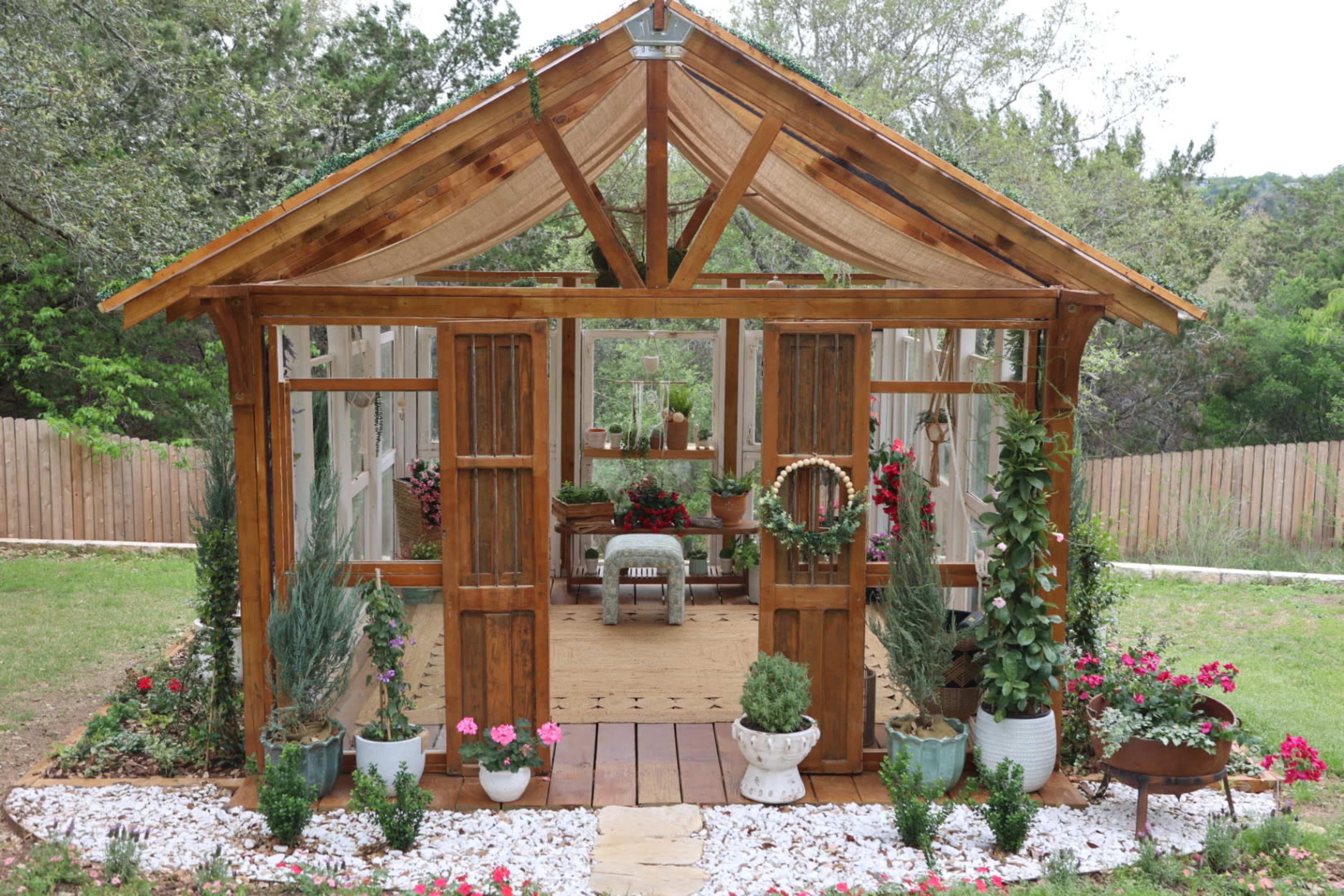 A wooden greenhouse with glass panels features flower pots and a seating area surrounded by greenery and flowers.
