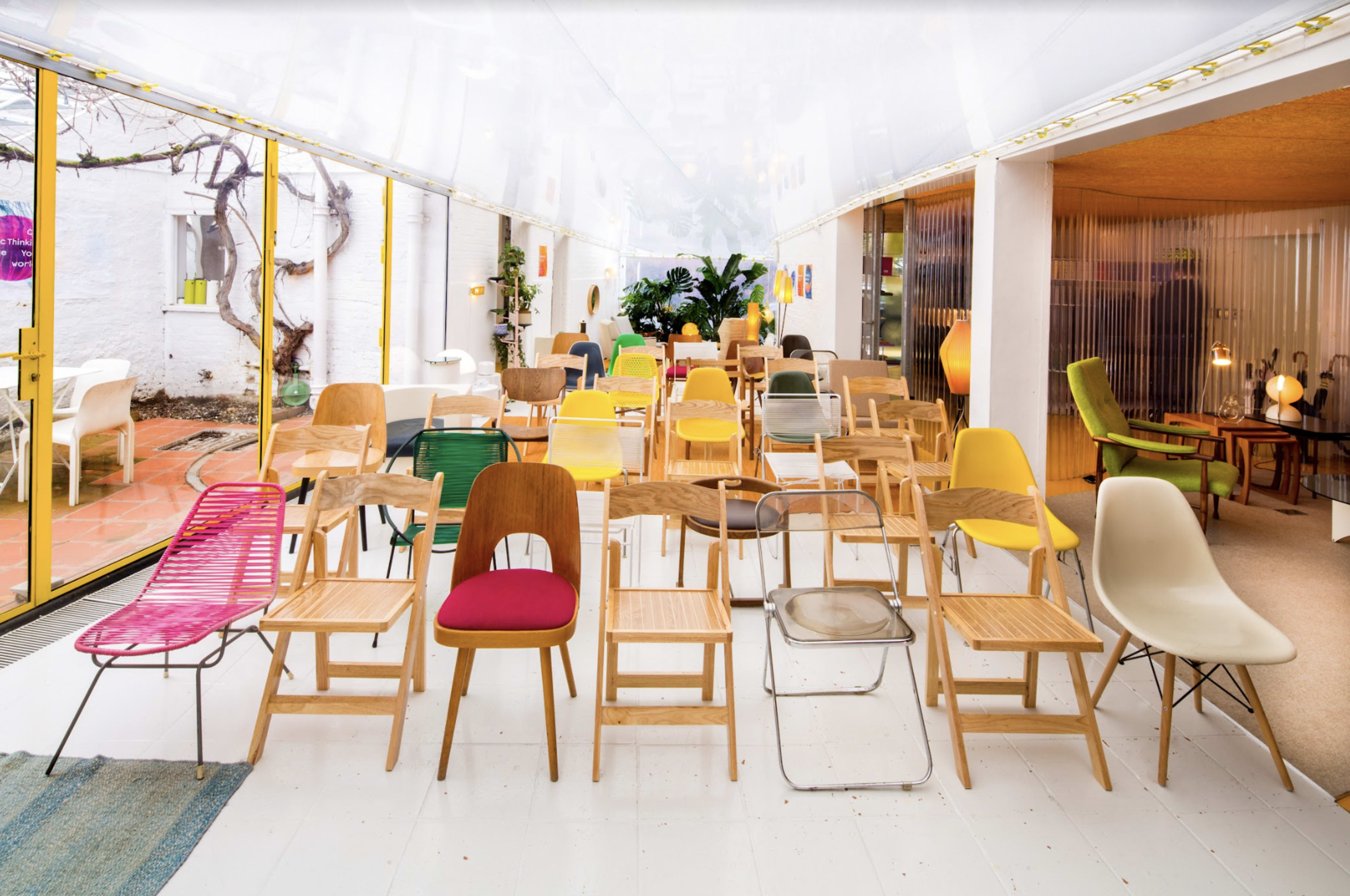 The image shows a spacious interior featuring an array of colorful chairs arranged in a line, with large windows allowing natural light to fill the room.