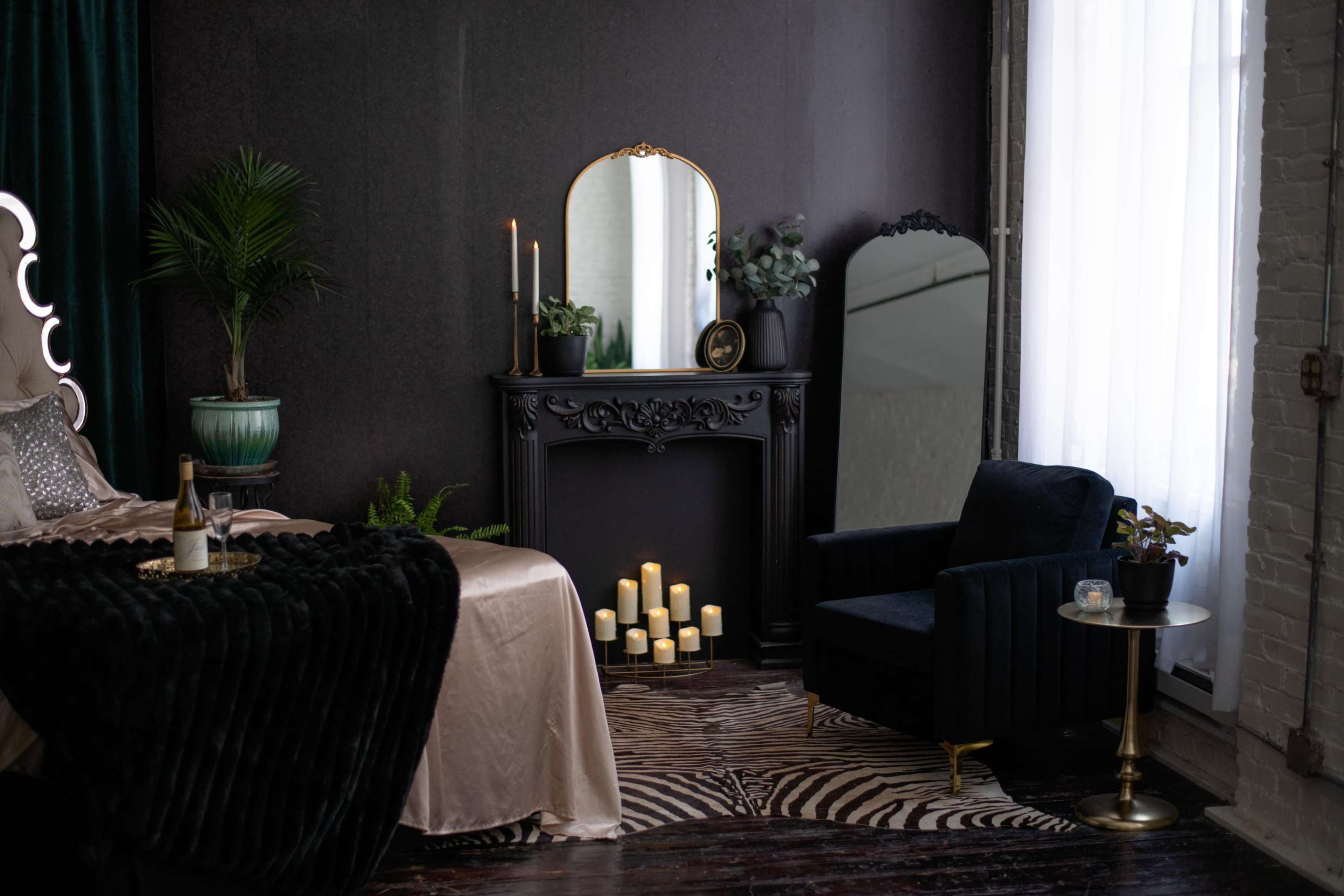 The image shows a stylish bedroom featuring a bed with dark bedding, a decorative fireplace with candles, a fuzzy rug, and an armchair beside a small table.