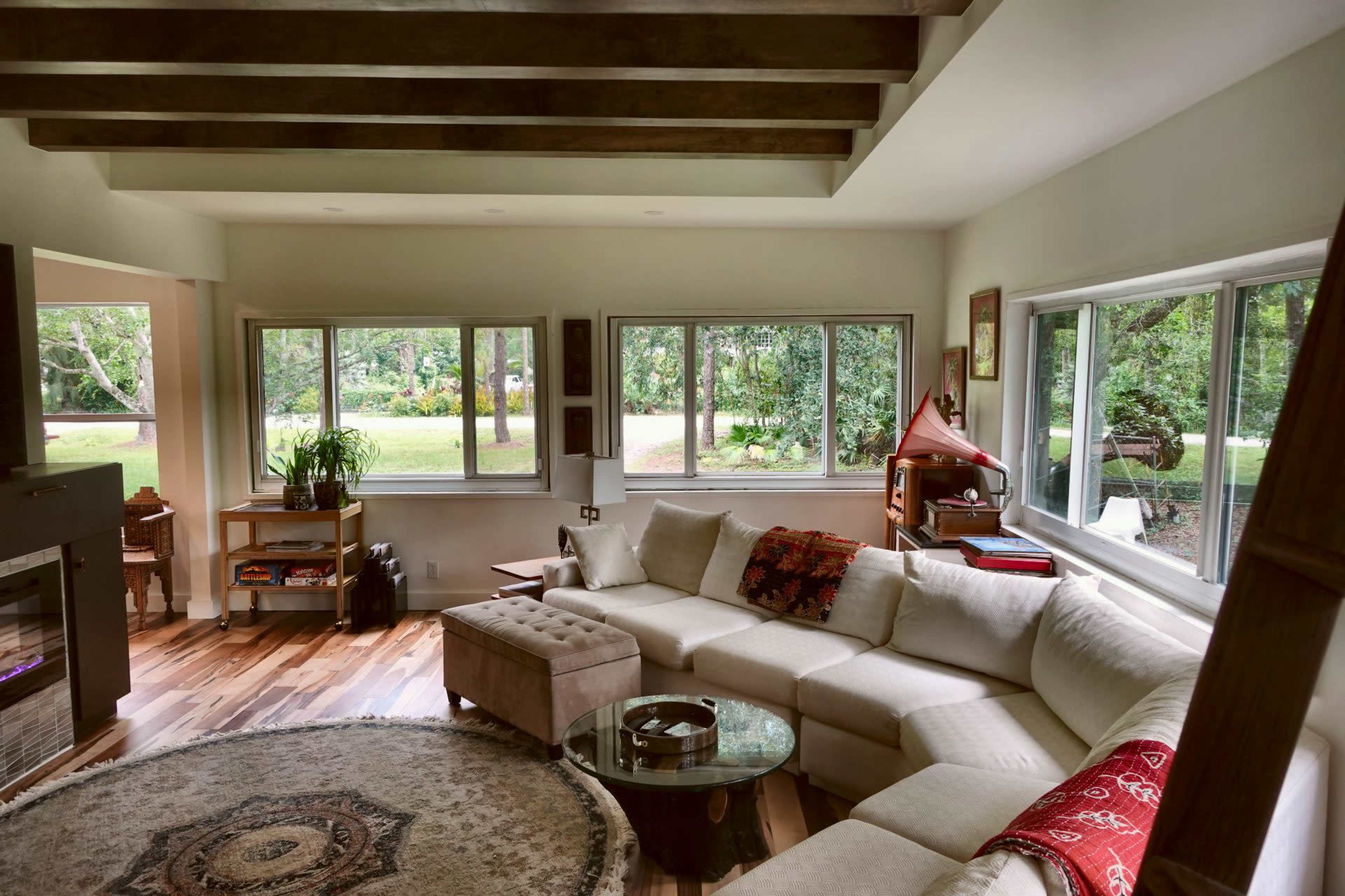 A spacious living room features a large sectional couch, a glass coffee table, and several windows providing views of an outdoor garden.