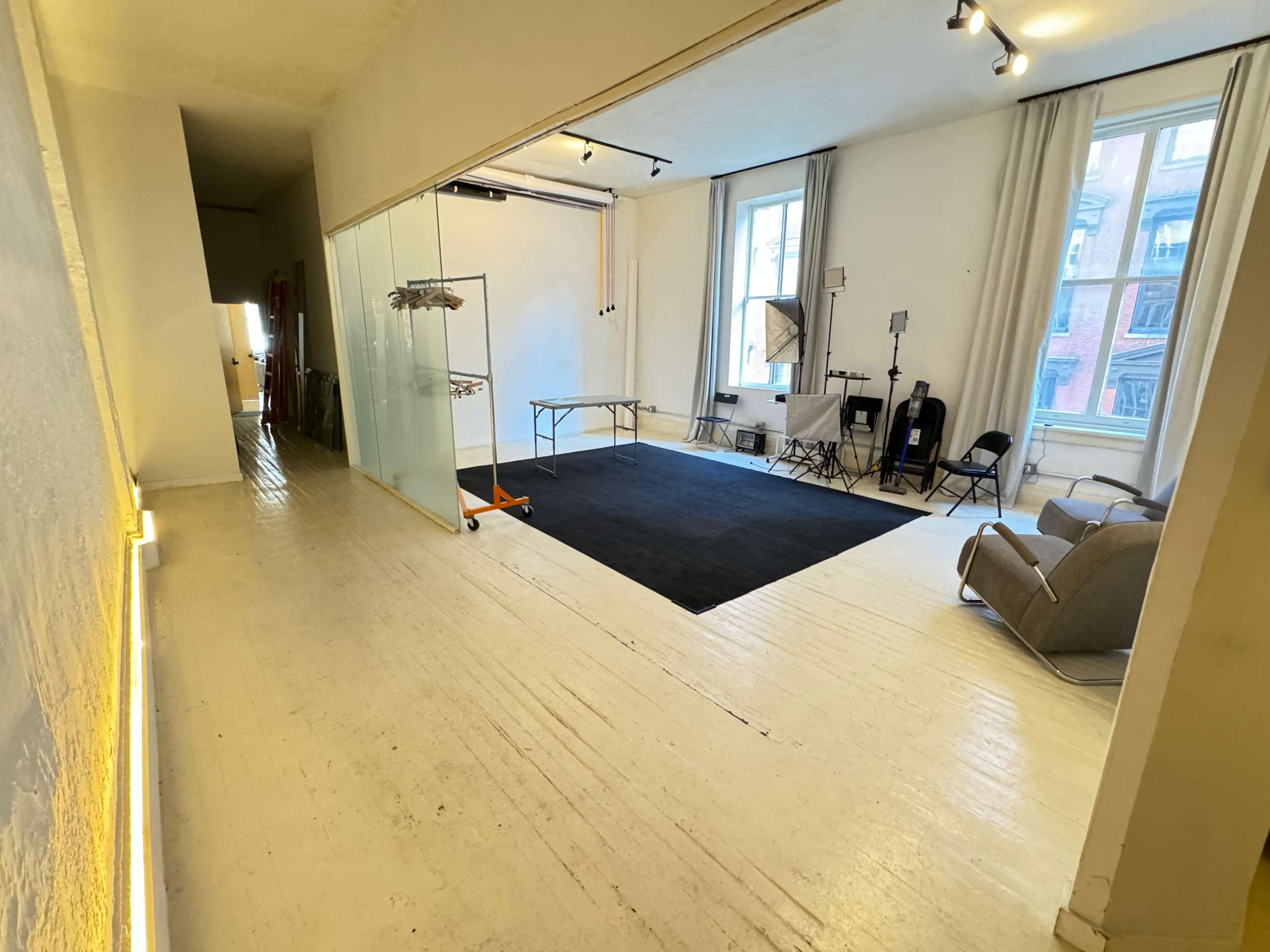 The image shows an indoor studio space featuring a large area with wooden flooring, a black carpet, a folding table, chairs, and lighting equipment.